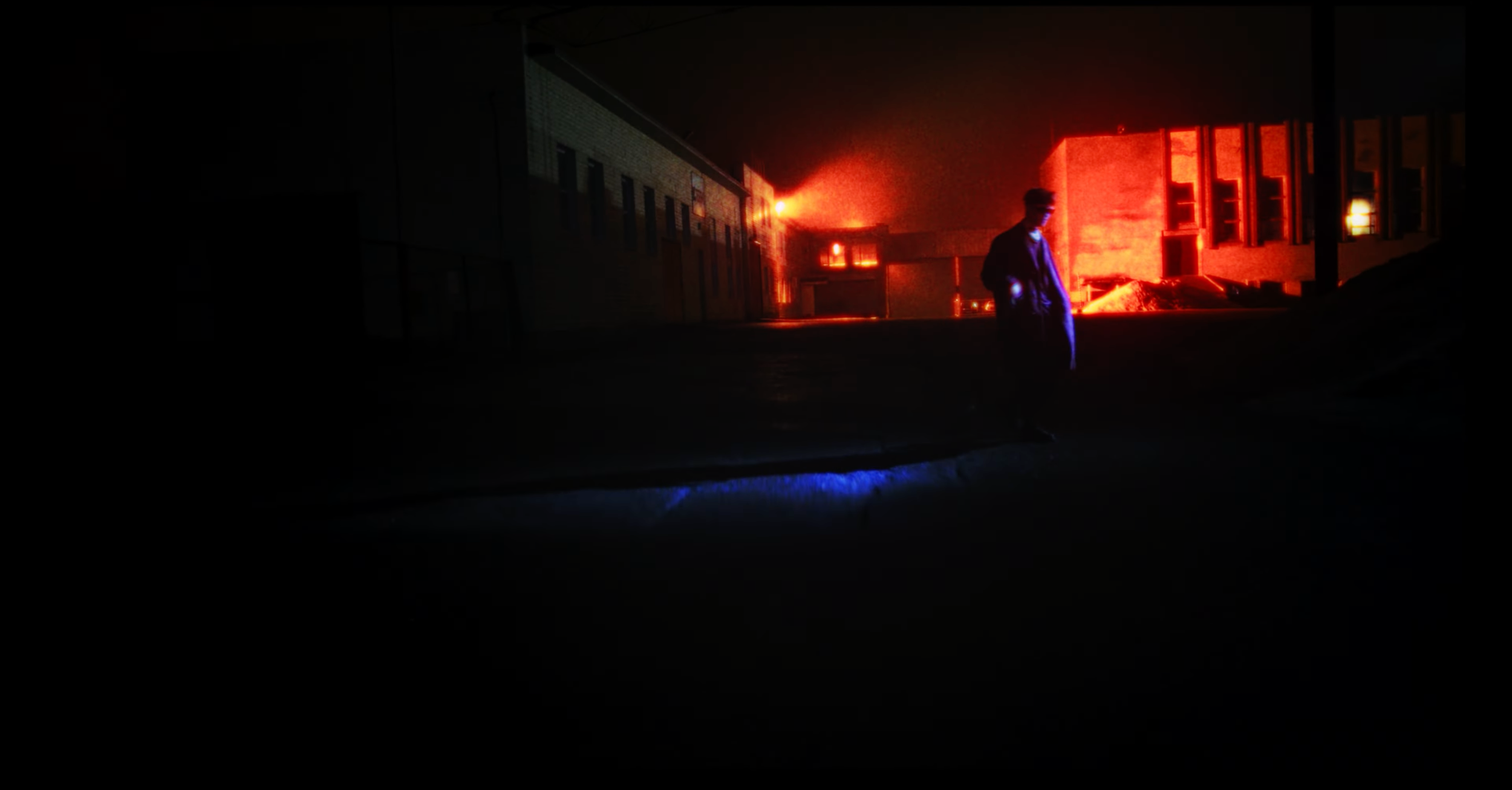 Nighttime scene of a person standing outside a building on fire, with glowing flames and smoke visible in the background.