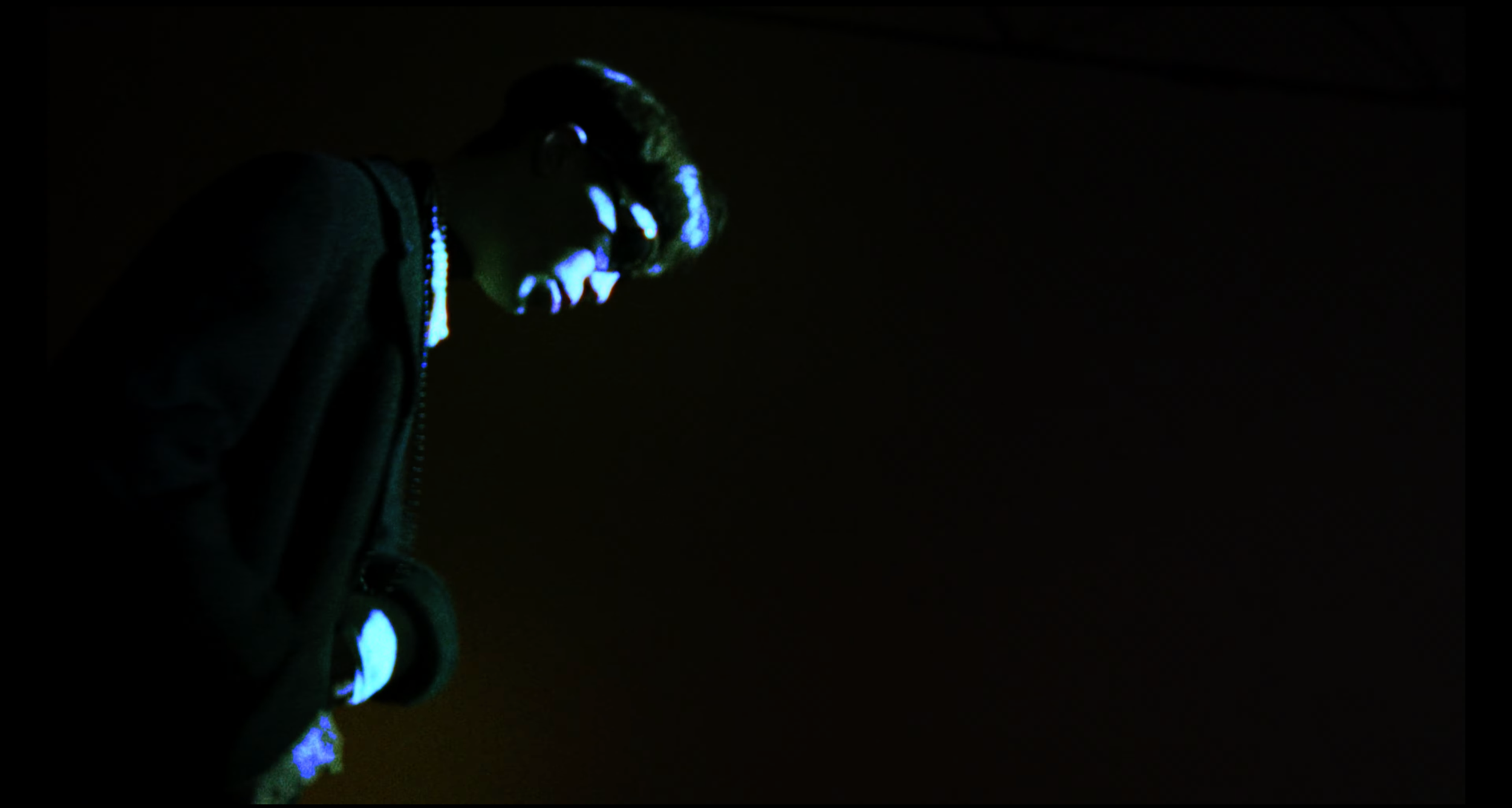 Person in dark clothing with glowing blue accents on their face and hand, in a dark environment.
