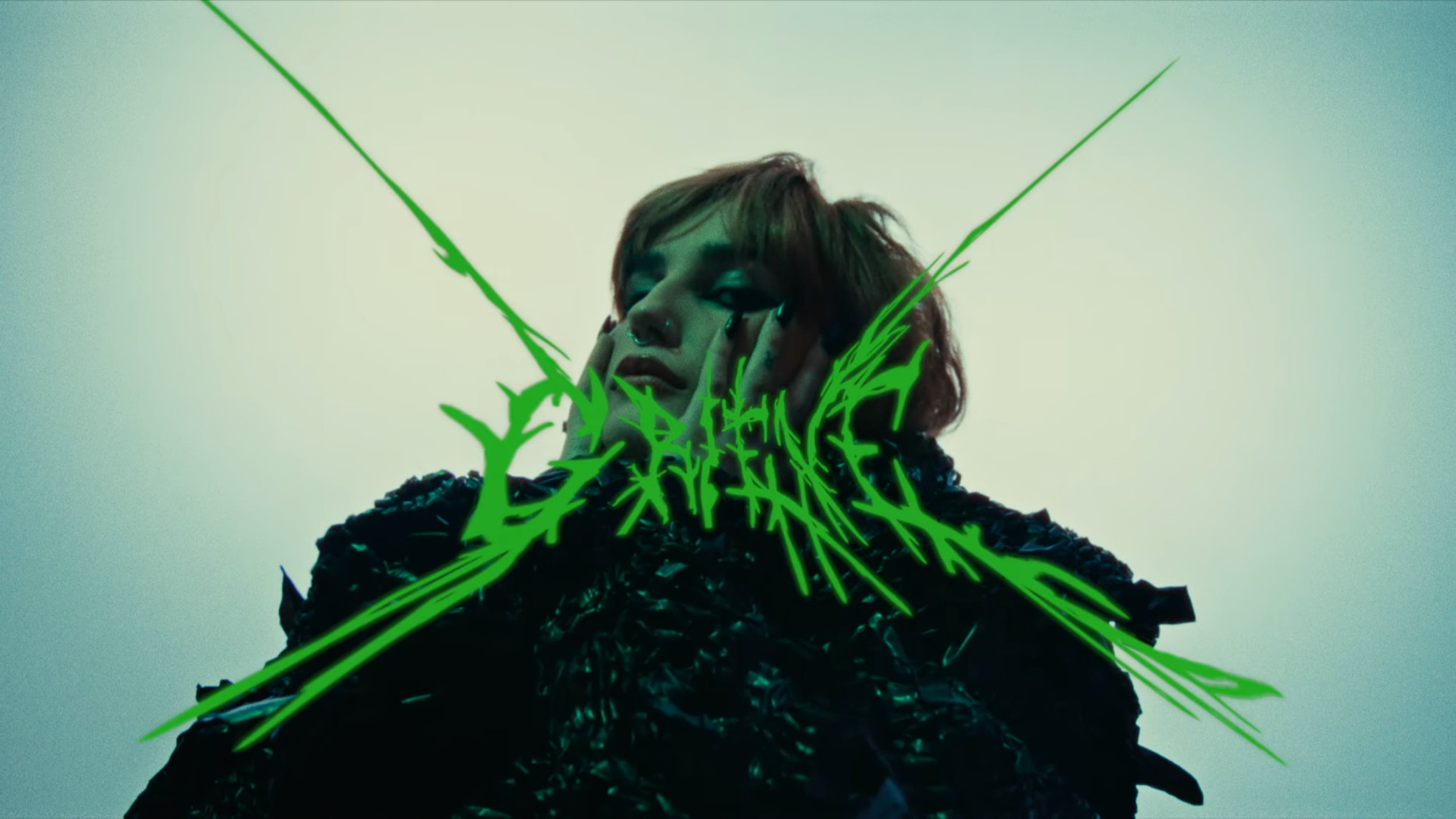 A person with reddish hair, makeup, and piercings, resting their face on their hands, with green graffiti-like text overlaying the image.