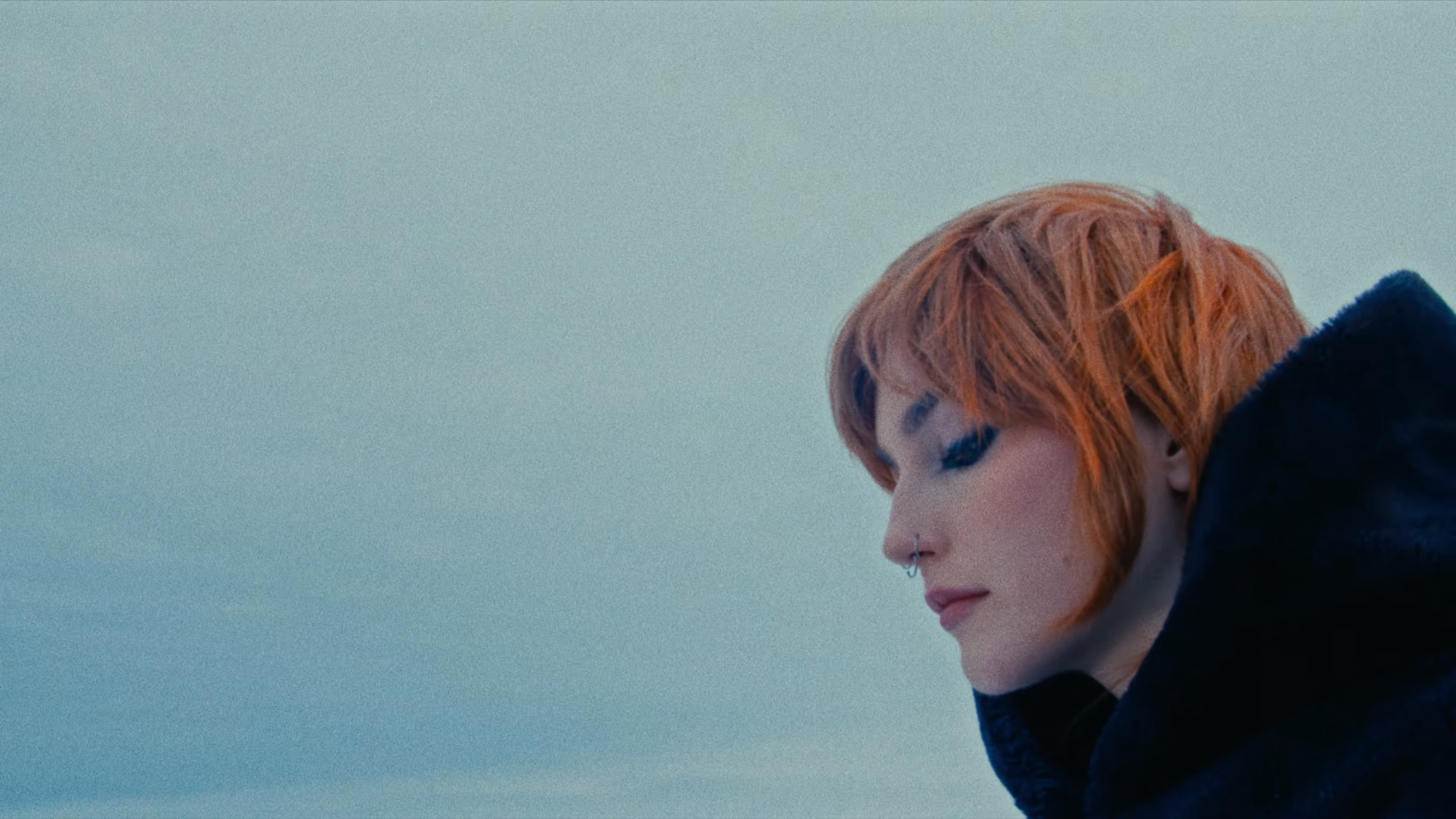 A close-up side profile of a person with short, reddish-orange hair, wearing a black hoodie, against a plain, light background.