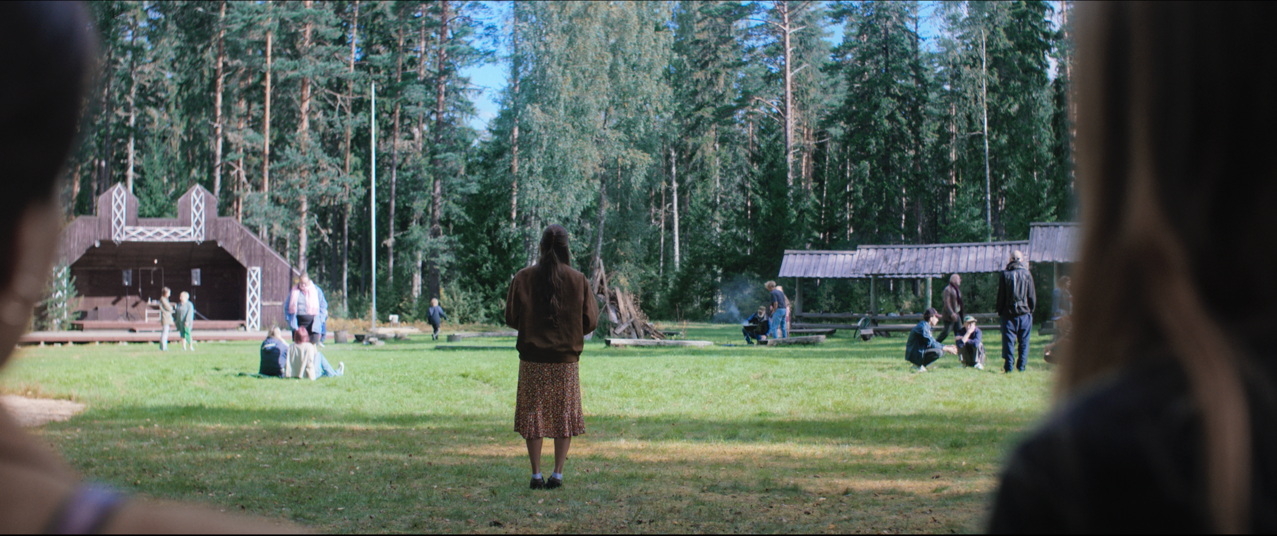 People gathered outdoors in a grassy park with trees in the background, some sitting or standing, and a woman standing in the center facing away from the camera.