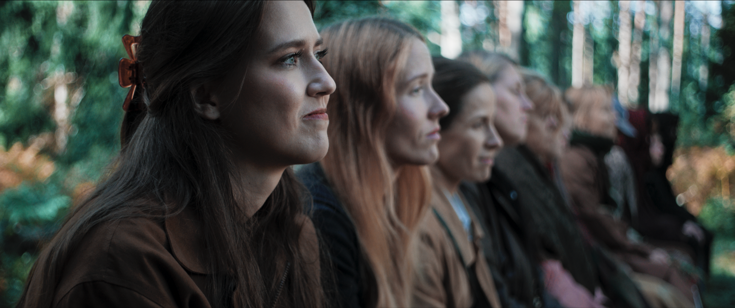 A group of women sitting outdoors in a line, looking attentively to the left, with trees in the background.