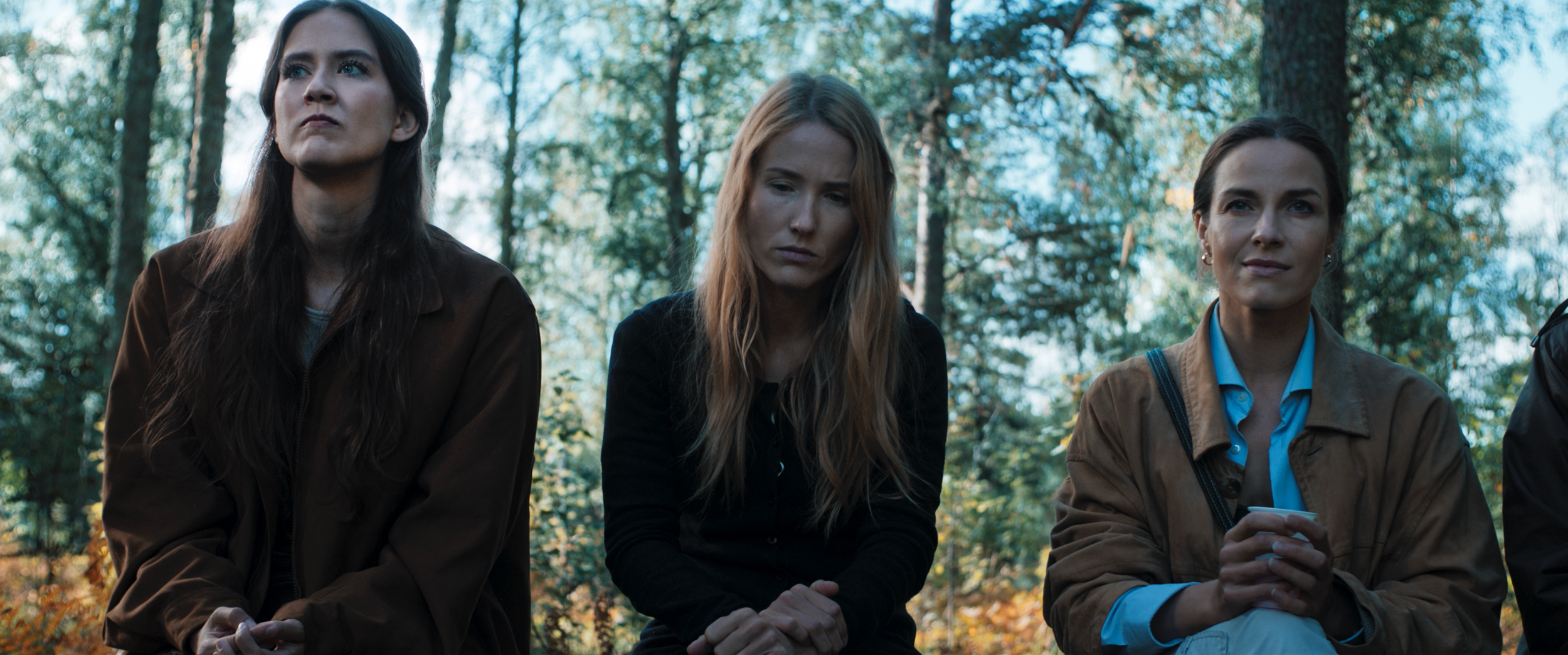 Three women sitting outdoors in a wooded area, appearing contemplative, with trees and sunlight in the background.