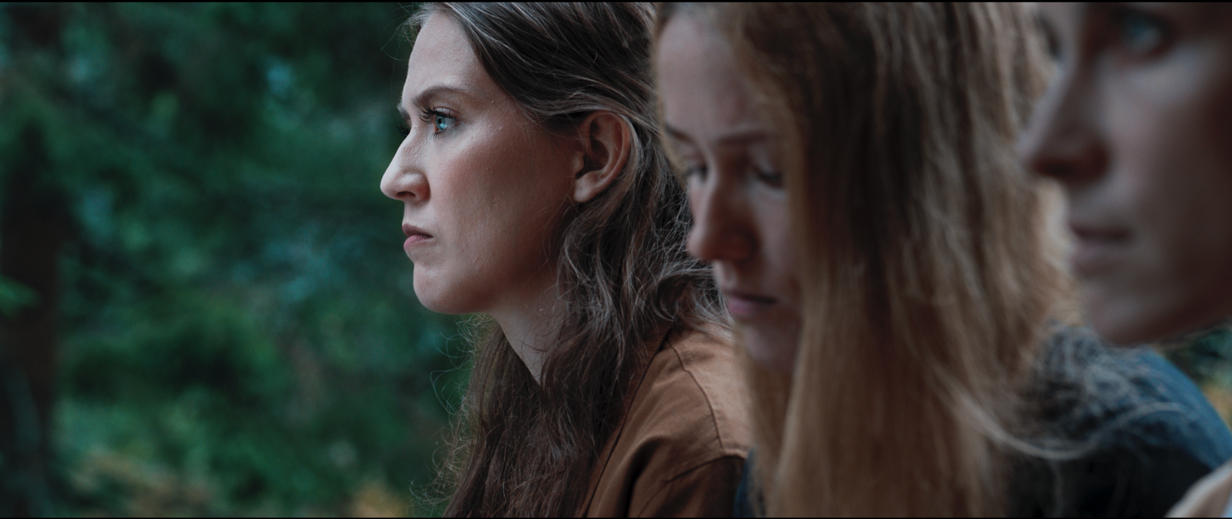 Three women sitting outdoors with trees in the background, the first woman in profile, the others partially visible, with thoughtful expressions.