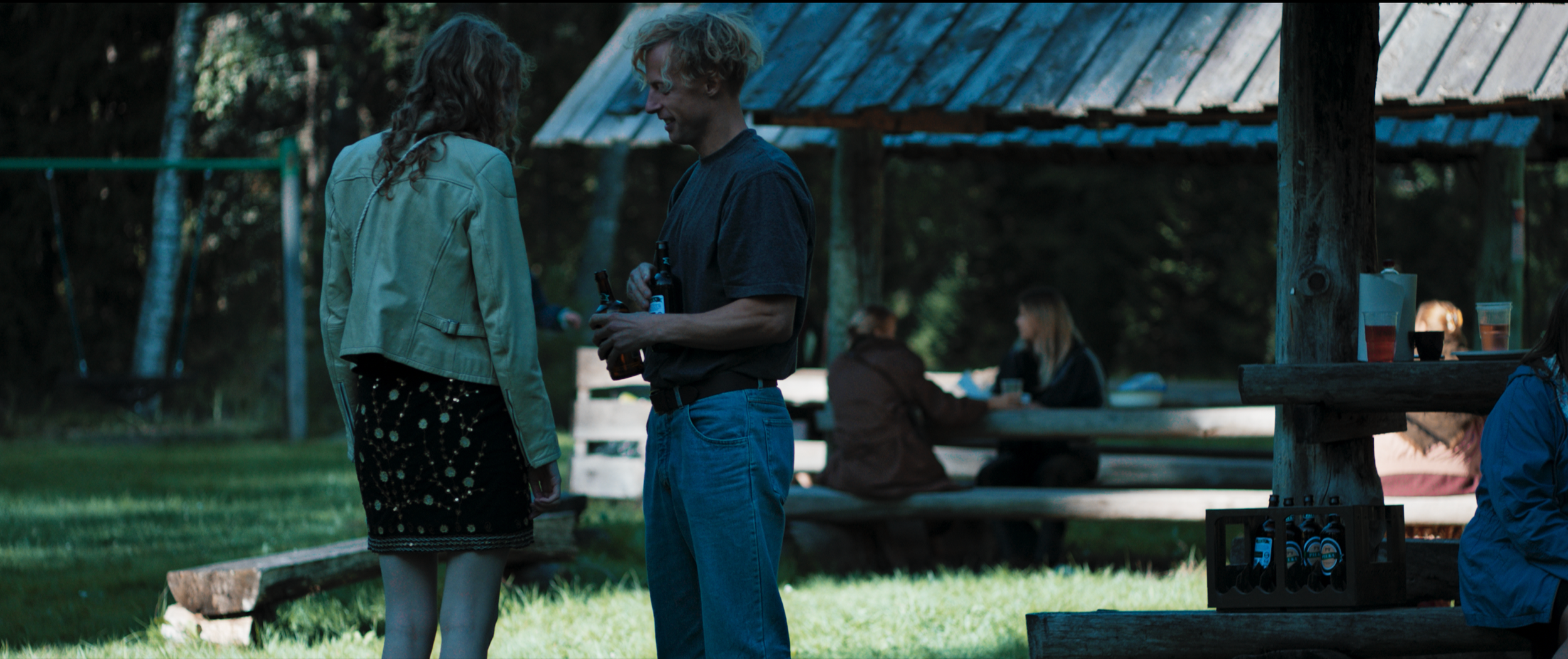 A man and woman stand close together holding bottles, chatting outdoors at a gathering, with a pavilion and other people in the background.