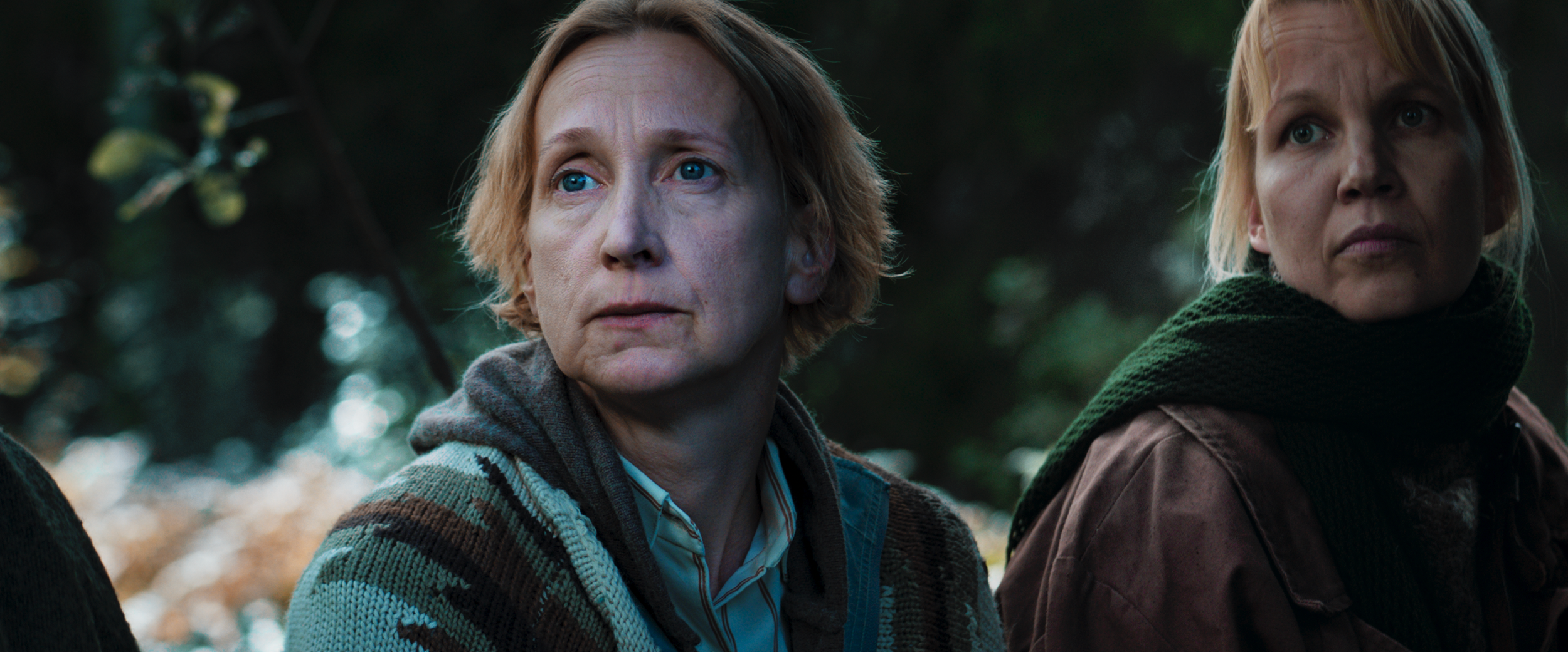 Two women sitting outdoors, looking thoughtfully into the distance with a blurred forest background.
