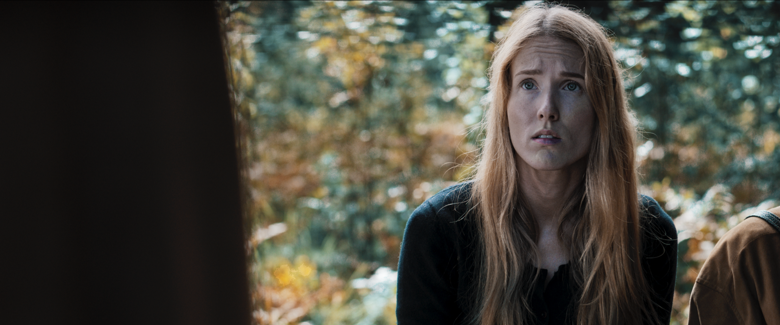 A woman with long red hair and a concerned expression, outdoors with blurred fall foliage in the background.