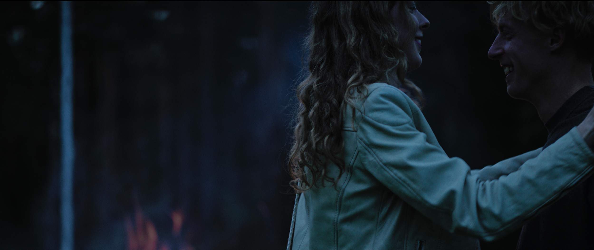 A couple holding each other, smiling and looking into each other's eyes in a dark, outdoor setting, possibly around a fire.