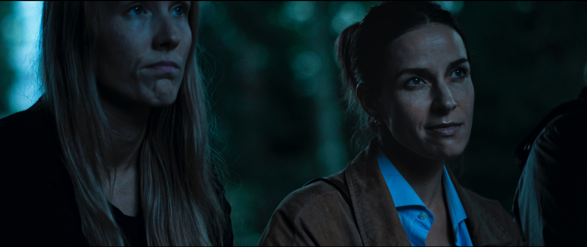 Close-up of two women outdoors, one with long blonde hair and the other with dark hair, looking attentively at something off-camera in a forest or park setting during the evening.