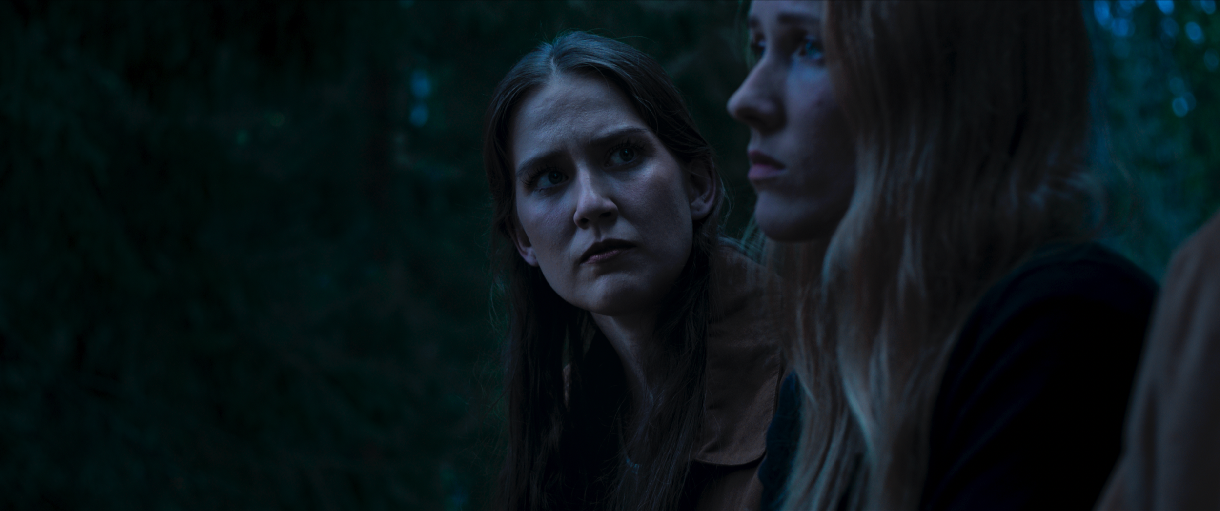 Two women with serious expressions in a dark, wooded setting, close-up shot.
