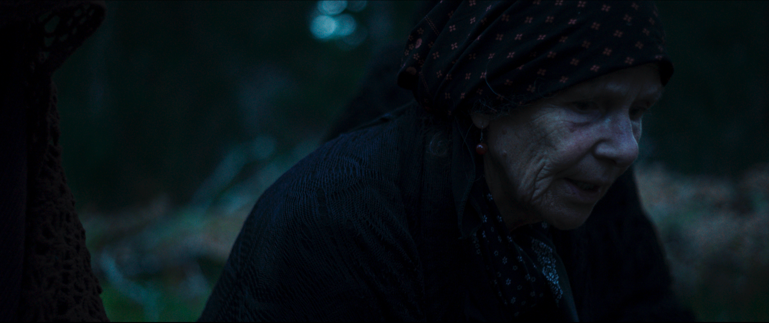 Close-up of an elderly woman with deep wrinkles, wearing a dark headscarf with red and white patterns and a black sweater, outdoors in a dimly lit environment.