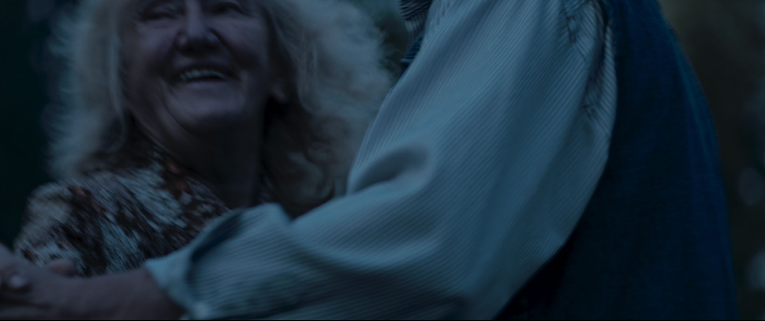 A person smiling and hugging another person outside during dusk or evening.