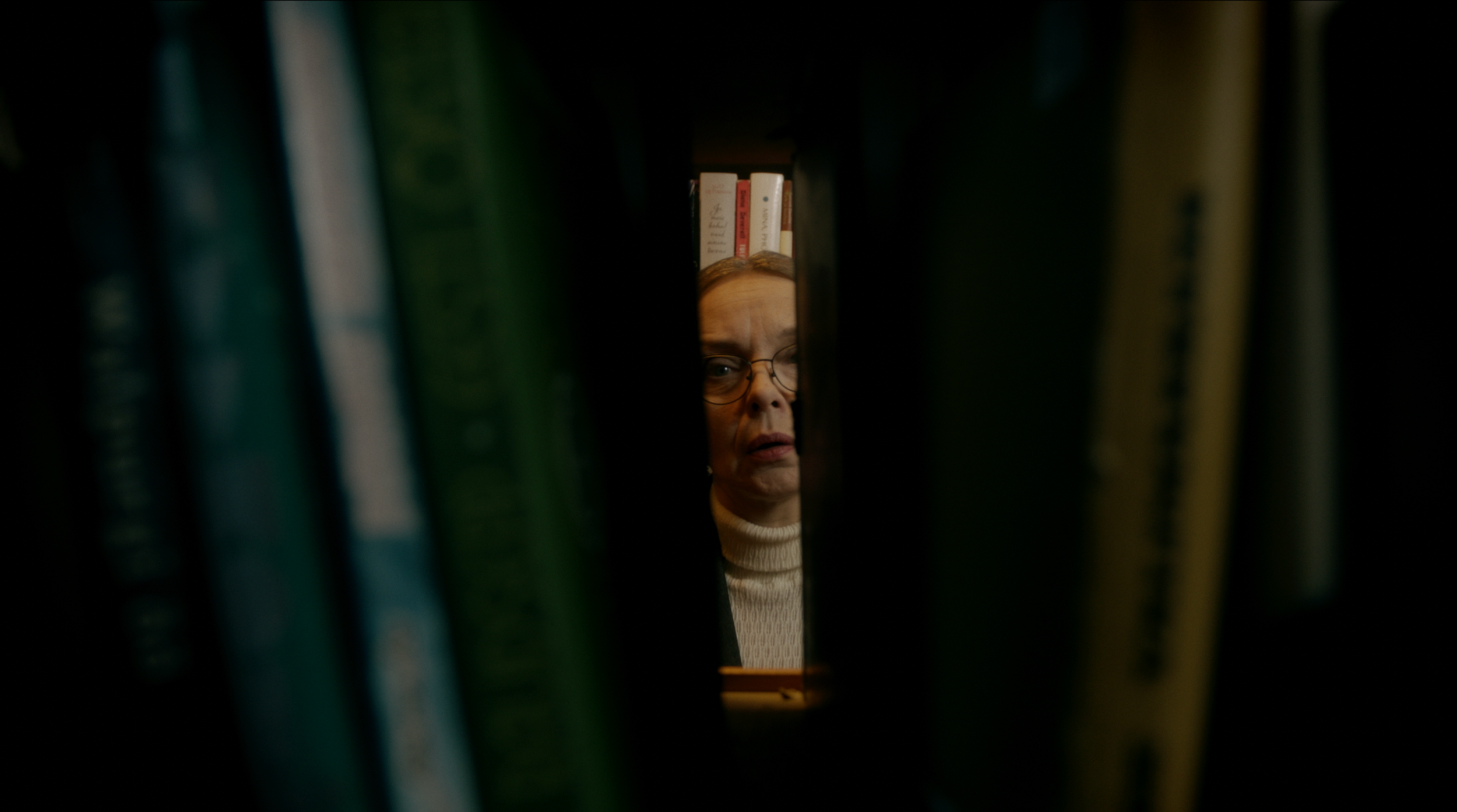 A woman with glasses looking through a narrow gap between books in a library.