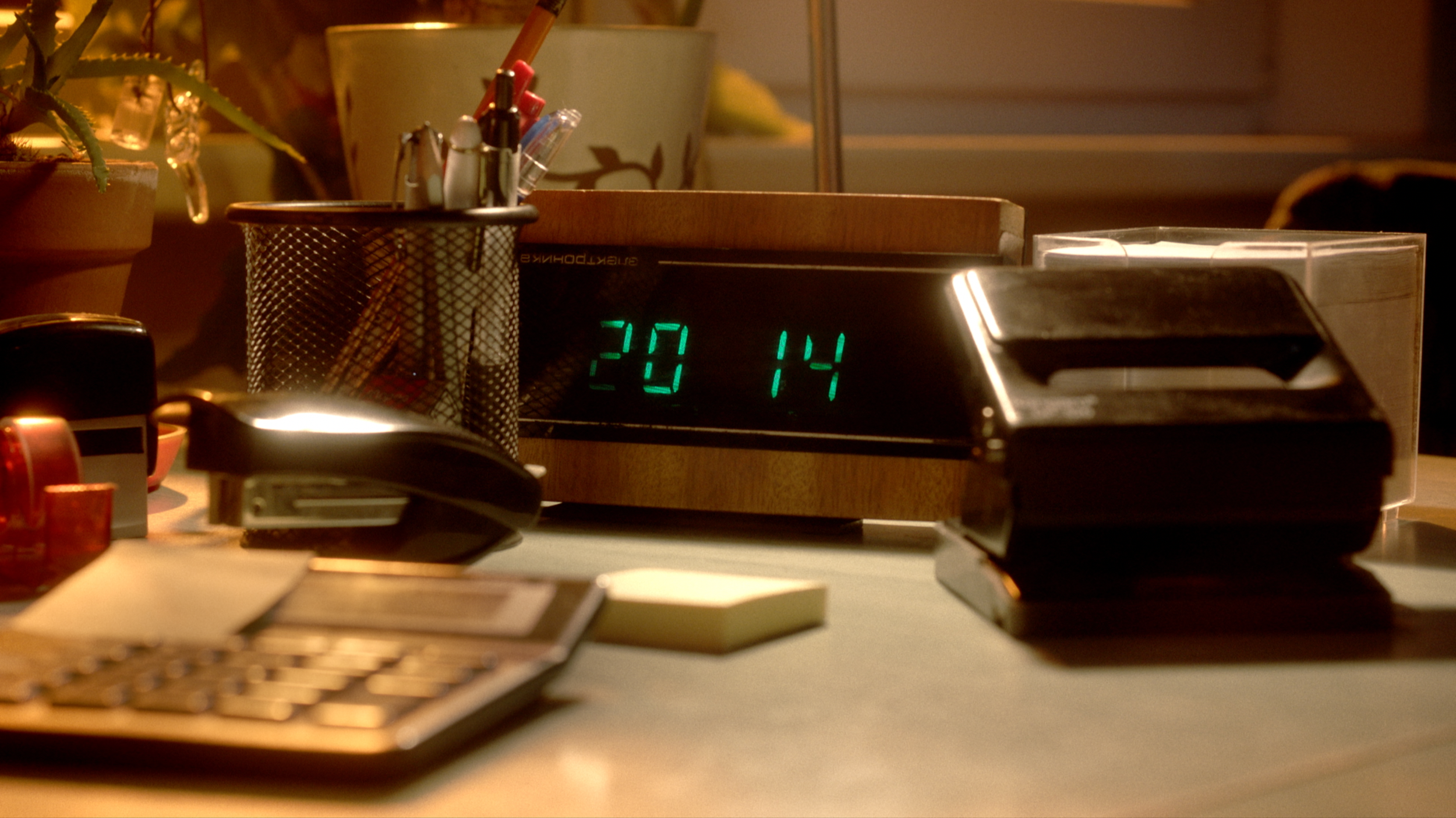 Close-up of a cluttered desk with a digital clock displaying 20:14, office supplies, a cordless phone, and a pen holder with pens, papers, and a small plant in the background.