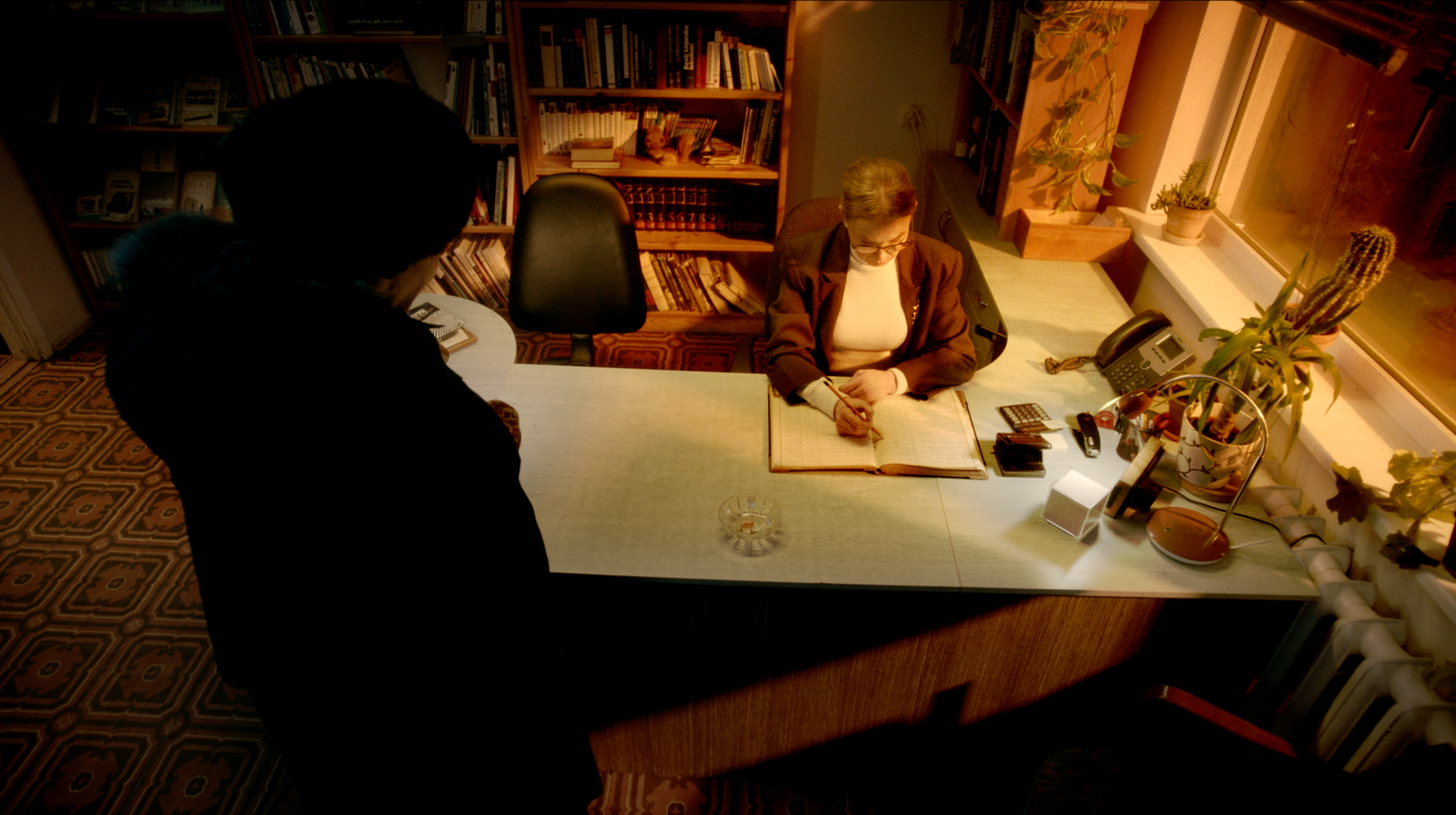 An older woman with glasses writing in a large open book at a desk, with a younger person standing nearby. The workspace includes a telephone, calculator, and various objects, with bookshelves behind and plants near a window.
