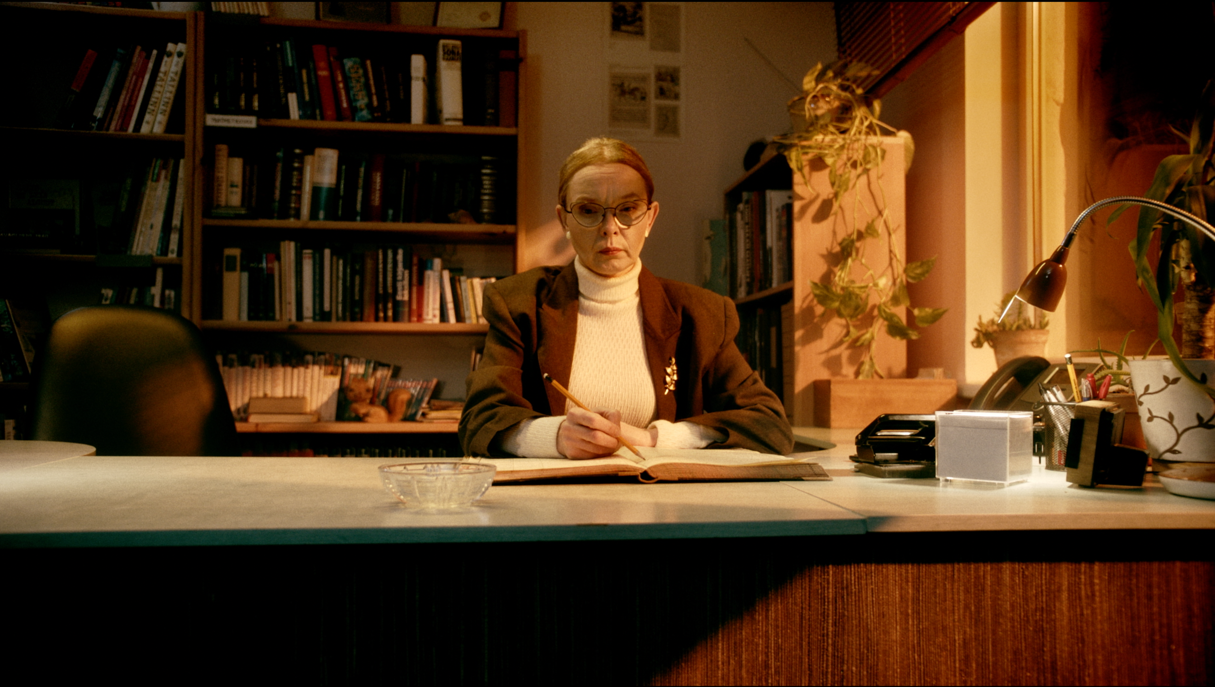 A woman with red hair and glasses seated at a desk, writing in a large book with a pencil, in a warmly lit room with bookshelves and plants.