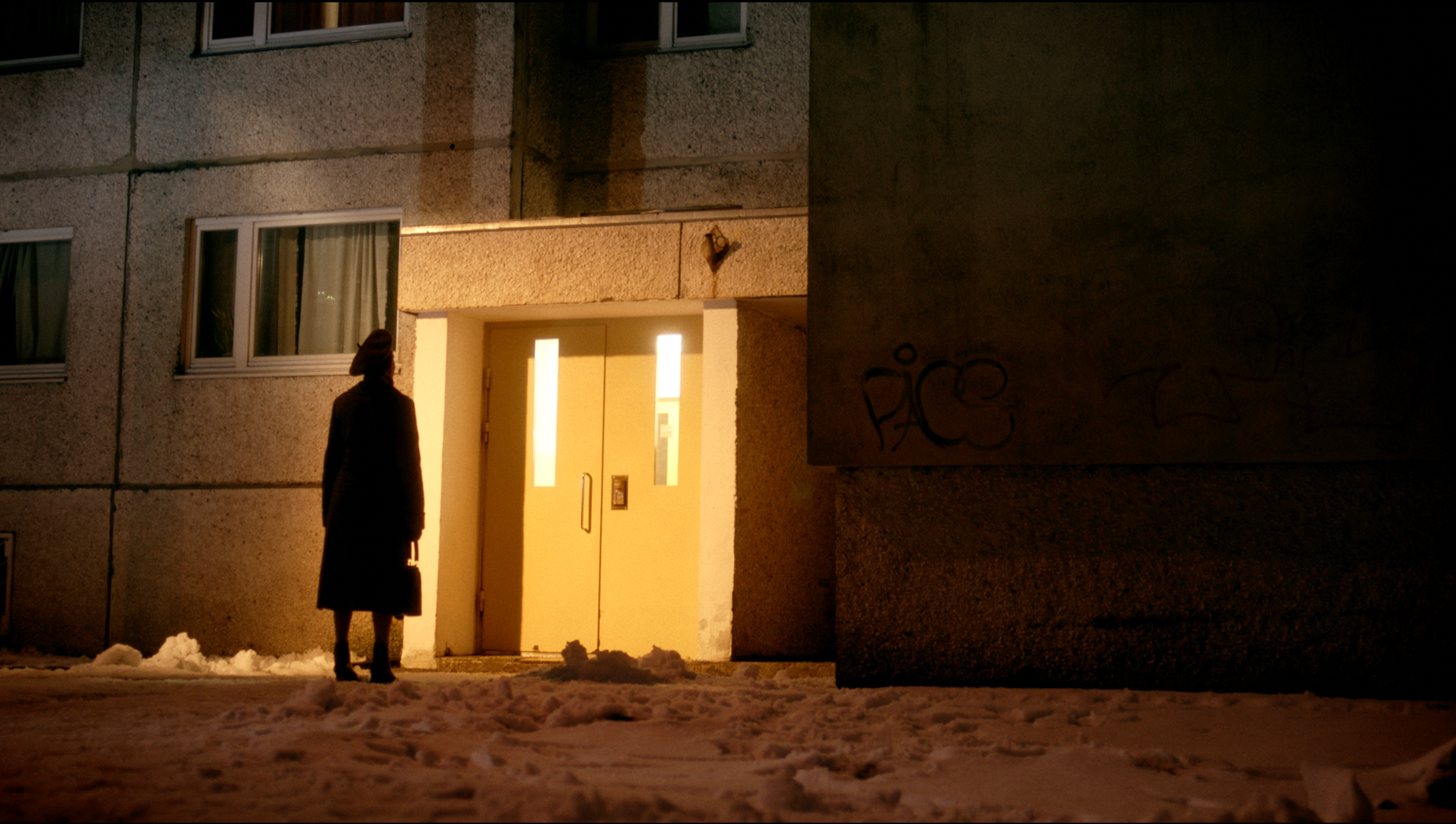 A woman standing outside a building at night, wearing a long coat and holding a bag, with snow on the ground. The building has illuminated windows and a lit entrance door.