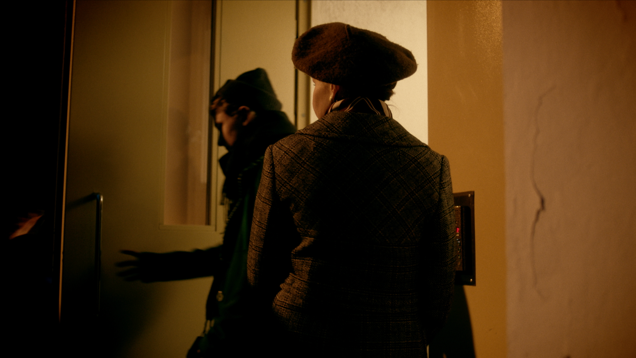 A woman wearing a brown beret and a plaid coat stands in front of a mirror in a dimly lit room, with a reflection of a young man in a beanie visible in the mirror.