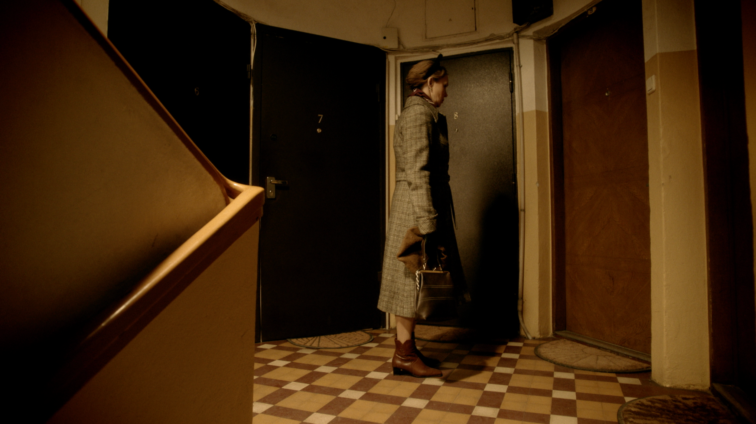 A woman standing in a hallway near a closed door, wearing glasses, a plaid coat, carrying a handbag and a coat, with her head slightly bowed.