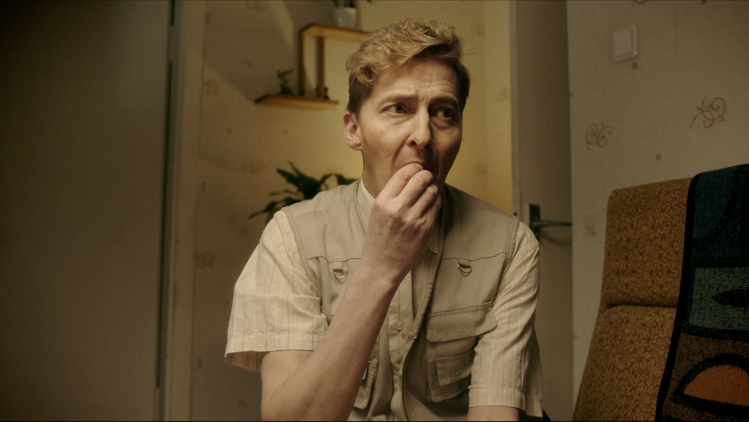 A person with light-colored hair and a beige shirt sitting at a table, touching their face with their hand, in a room with patterned wallpaper and a chair with a red and black patterned upholstery.