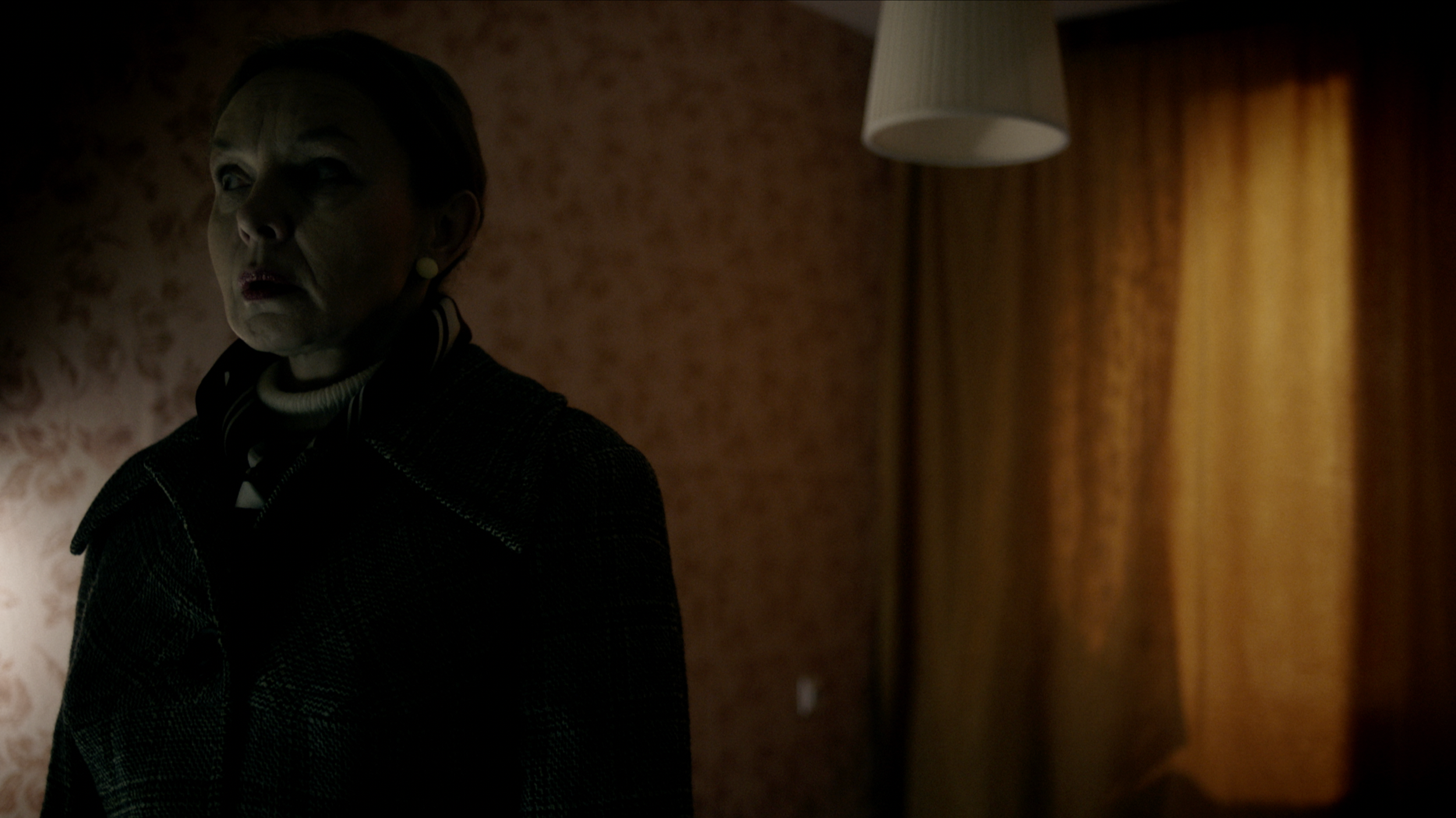 Woman with short hair and earrings in a dimly lit room with patterned wallpaper and curtains.