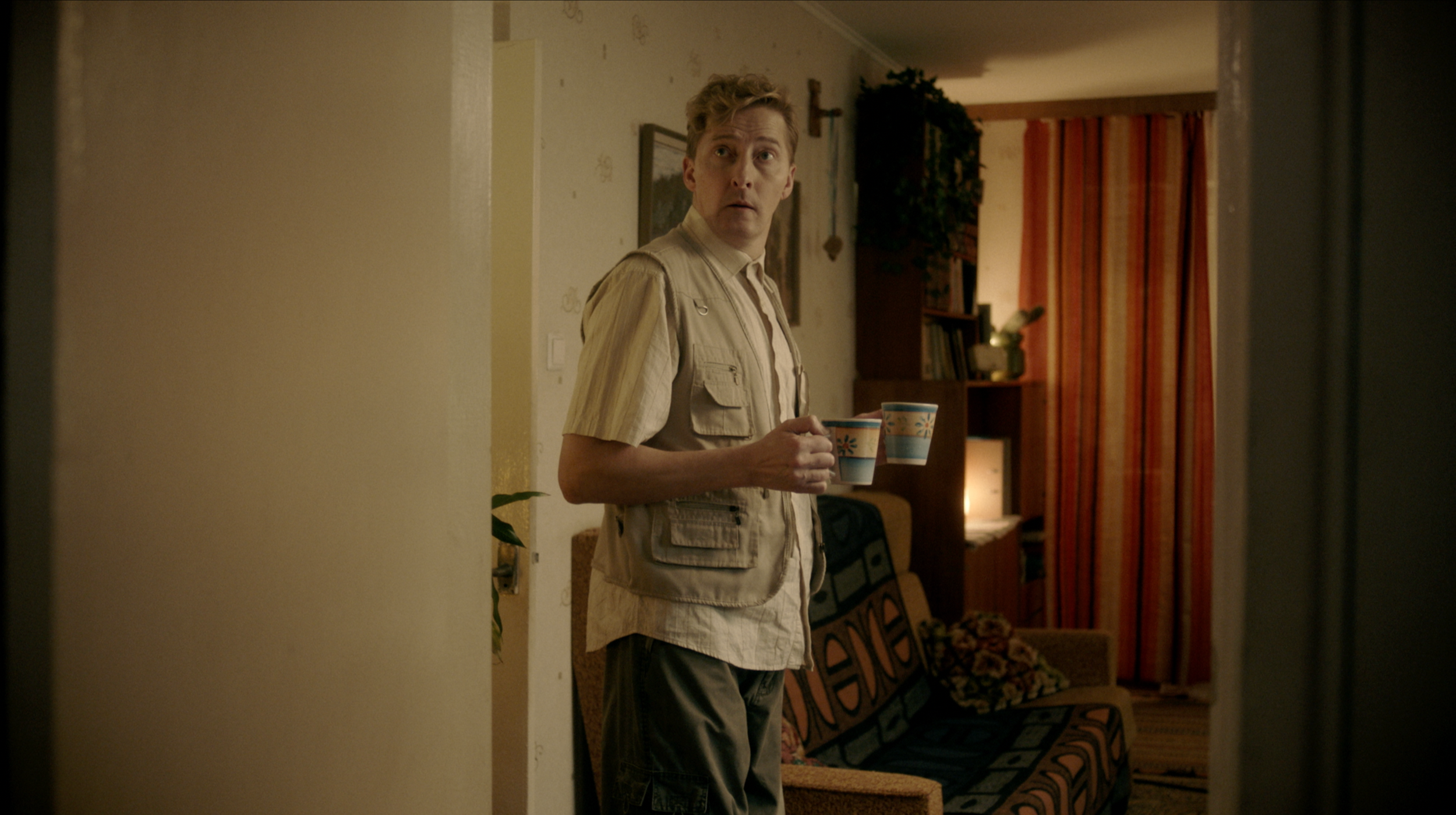 A man standing in a living room holding two cups, one in each hand, looking to the side with a surprised expression. The room has a couch with a patterned cover, shelves with books, and curtains in the background.