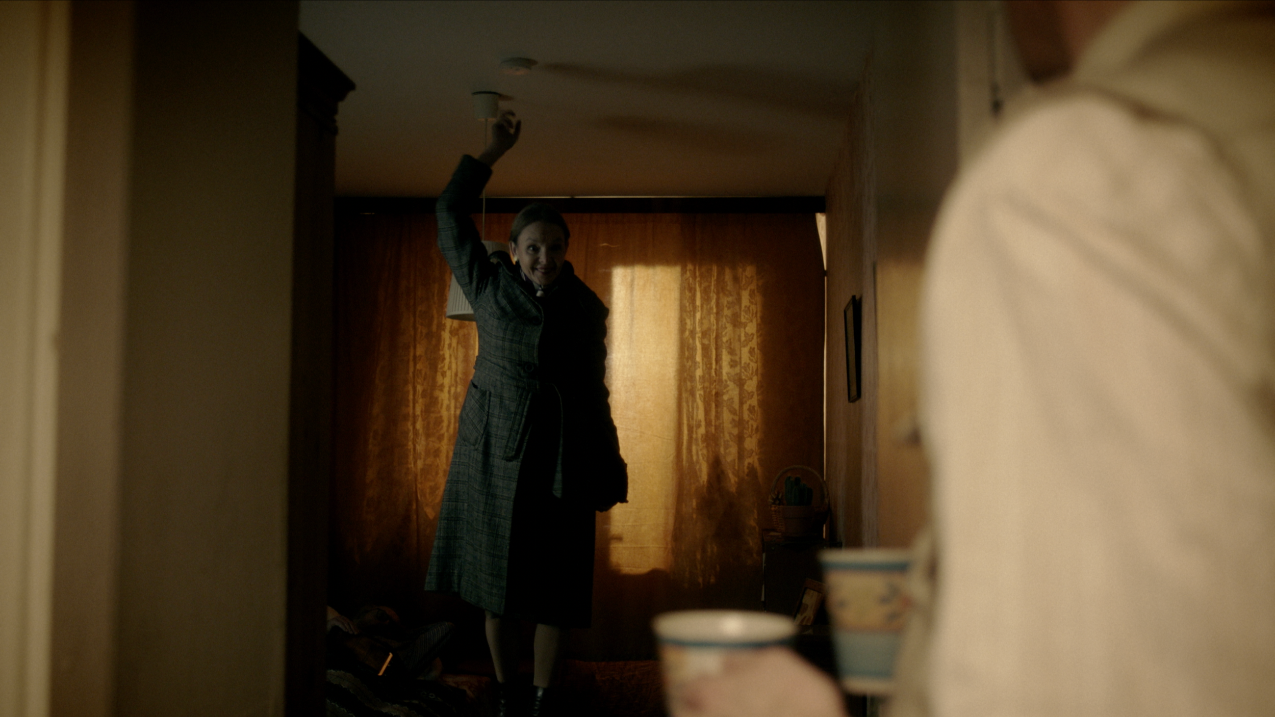 A woman in pajamas smiling and holding a smoke detector in an upper corner of a room, viewed from a low angle.