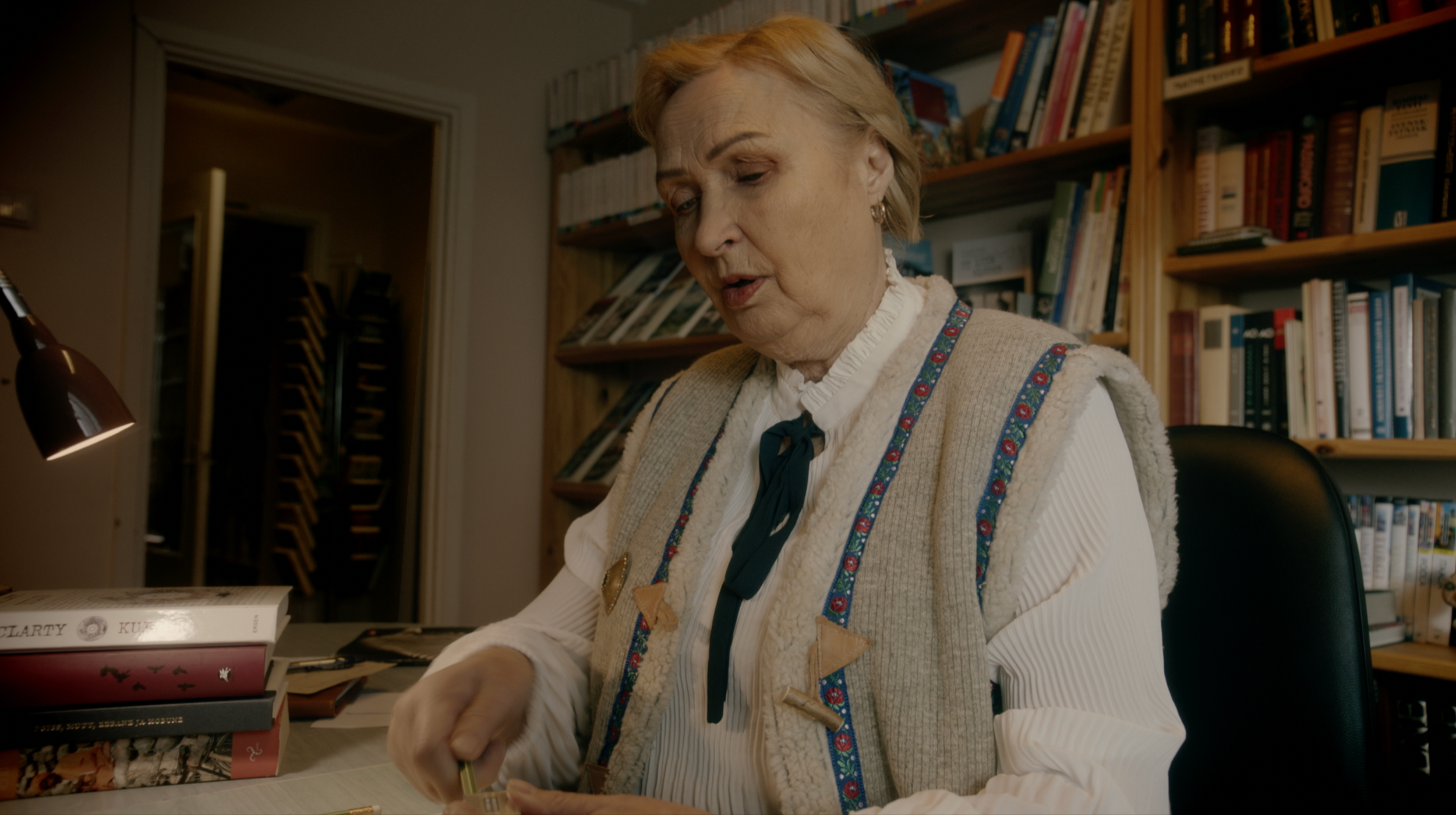 An elderly woman with blonde hair, wearing a beige vest with decorative trim, is sitting at a desk in a room filled with bookshelves. She is looking down, focusing on a book or paper in front of her.