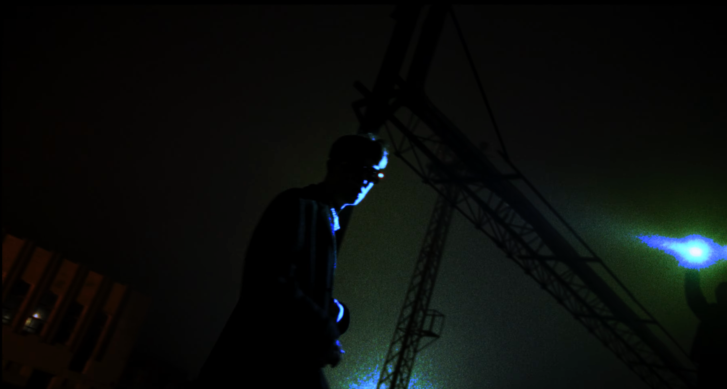 Person wearing a helmet and headlamp looking at the night sky with a laser beam pointing toward a structure, illuminated by a bright light source in the background.