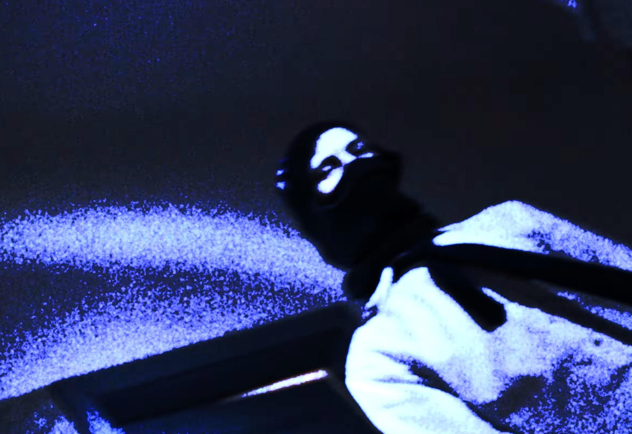 Person wearing black clothing with a skull print on the hood, spraying a fine white powder in a dark environment illuminated with blue light.