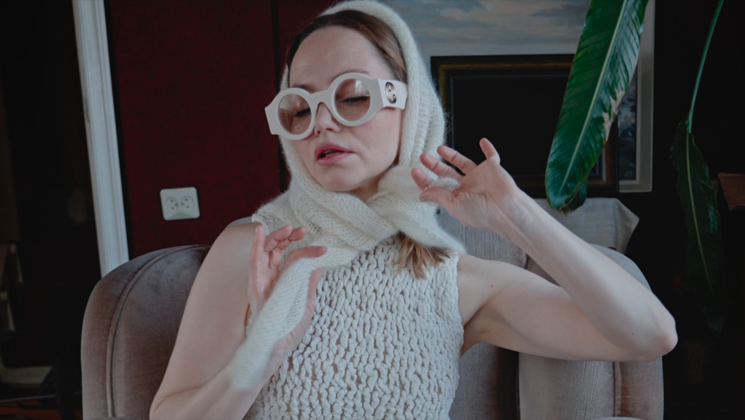 A woman wearing large white glasses, a white wool scarf, and a sleeveless textured sweater, sitting on a gray armchair in a room with a red wall, a black framed TV, a large green leafy plant, and a white electrical outlet.
