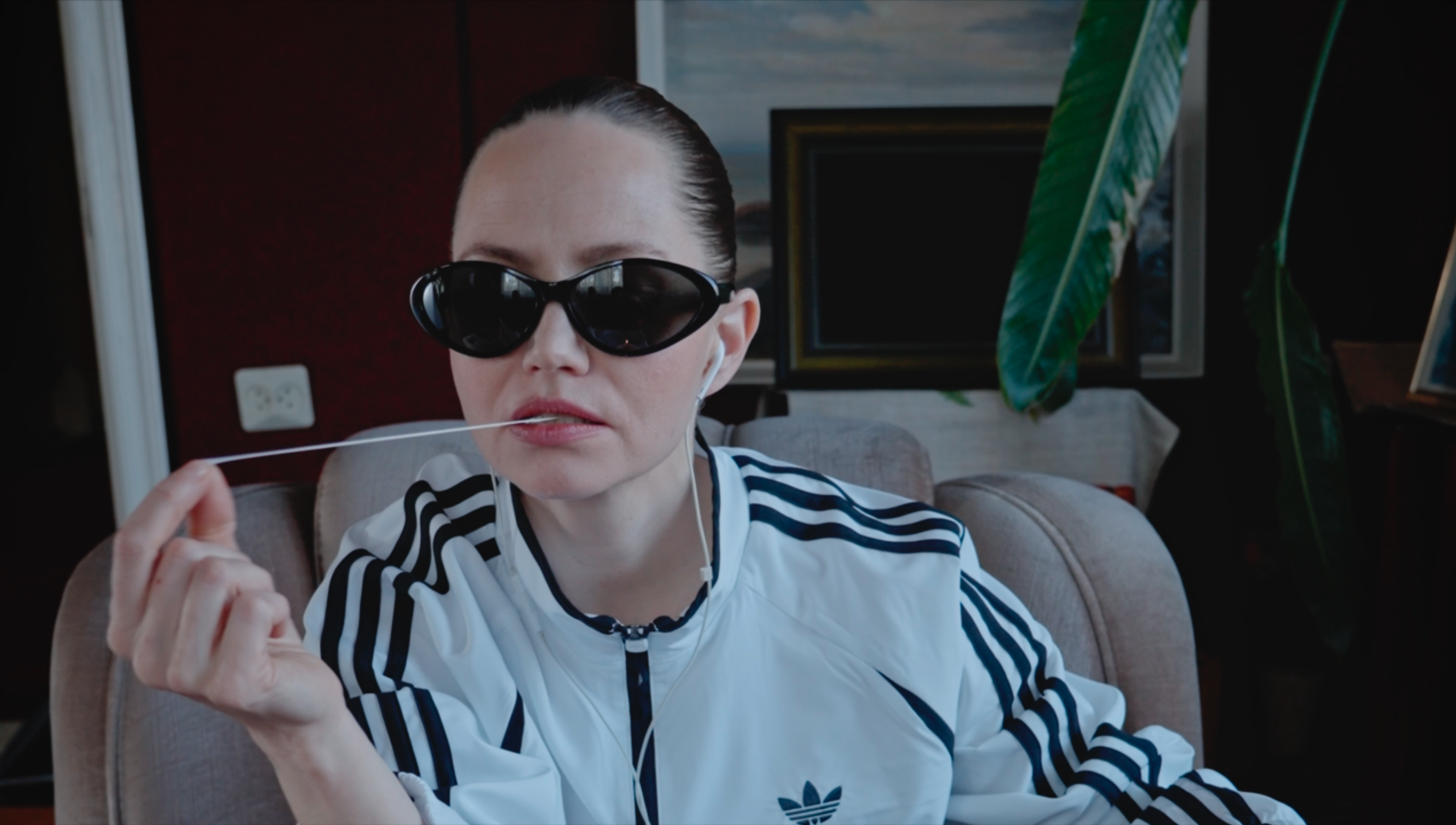 A woman with slicked-back hair wearing black sunglasses, a white sports jacket with black stripes, and earbuds, is sitting on a beige chair in a room with plants and framed pictures in the background.