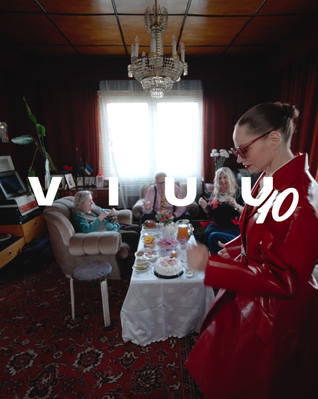 A woman in a red leather jacket and glasses smiling at elderly women sitting on couches in a living room. There is a table with a cake and drinks, a chandelier hanging from the ceiling, and the word 'ViiU Yo' in white text over the image.