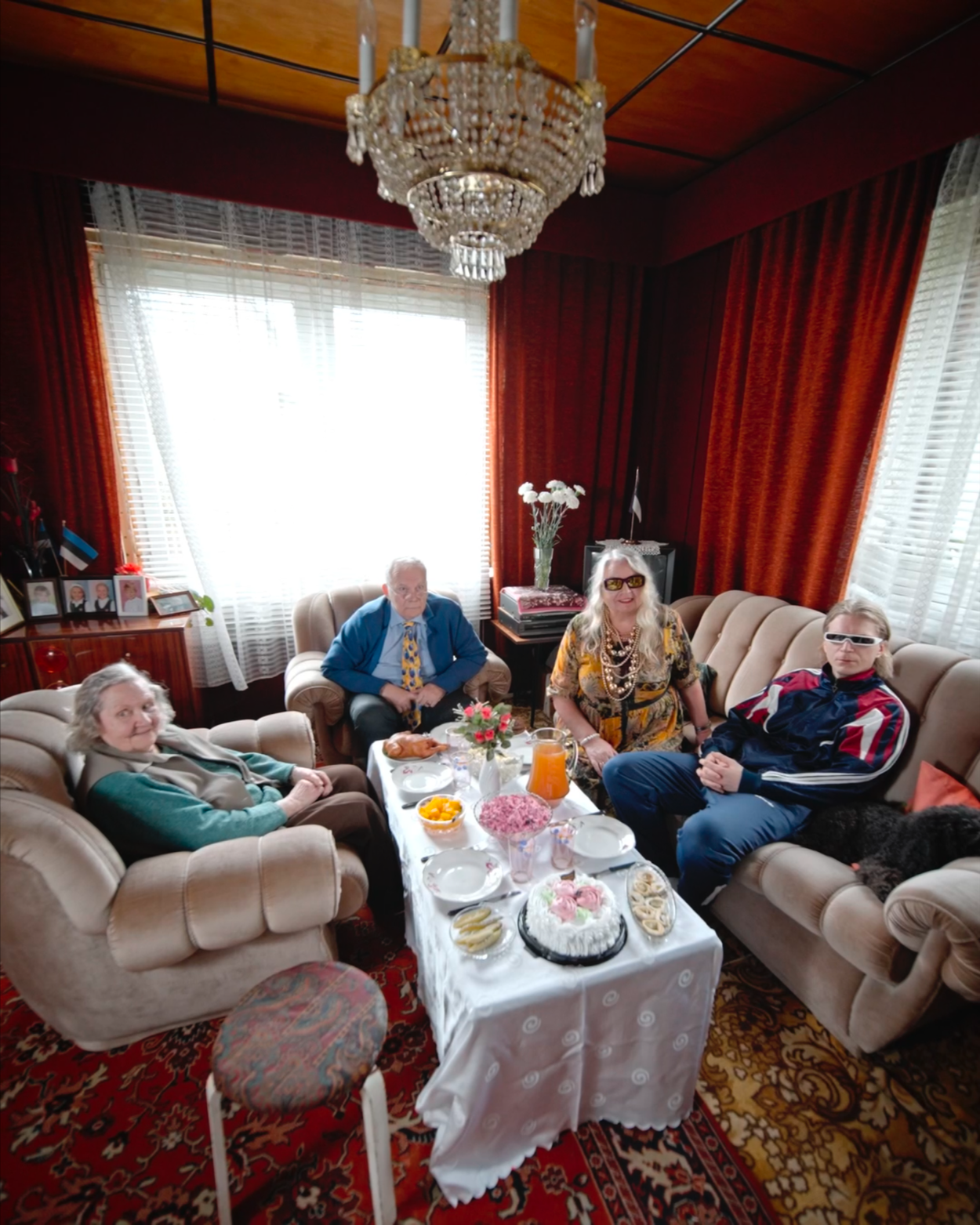 Four people sitting in a living room with a table of food and drinks, red curtains, and a chandelier overhead.