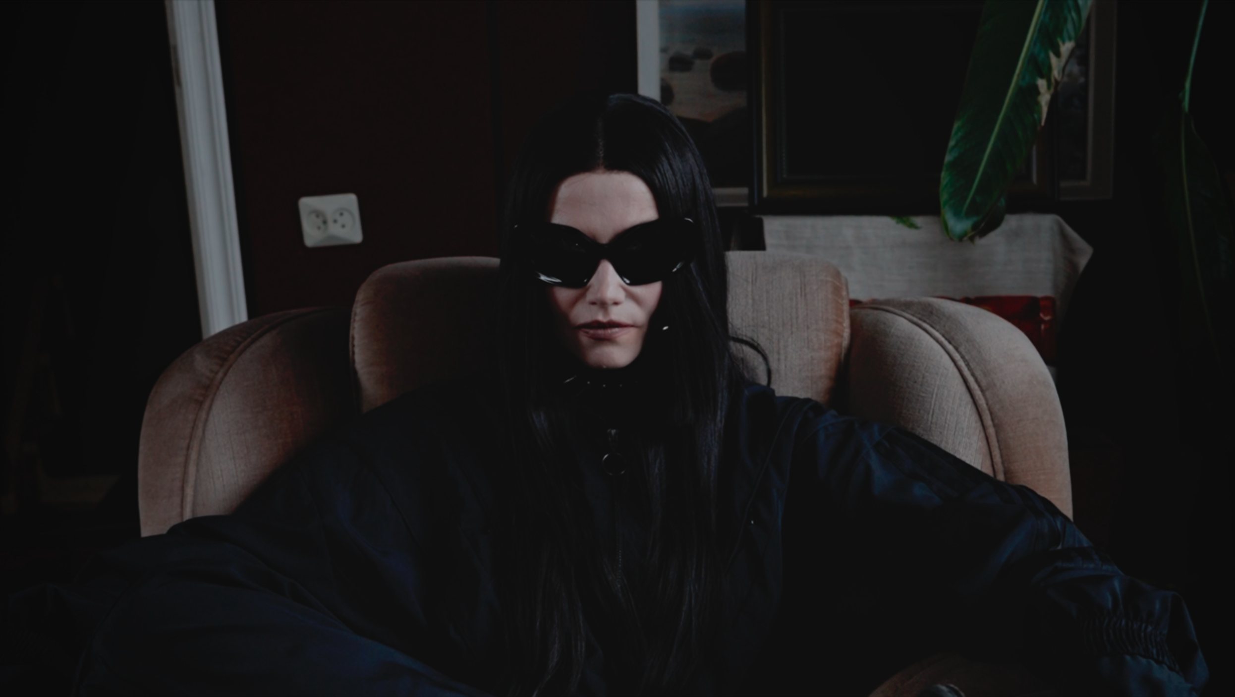 Woman with long black hair wearing black sunglasses and a black jacket sitting on a beige armchair indoors.