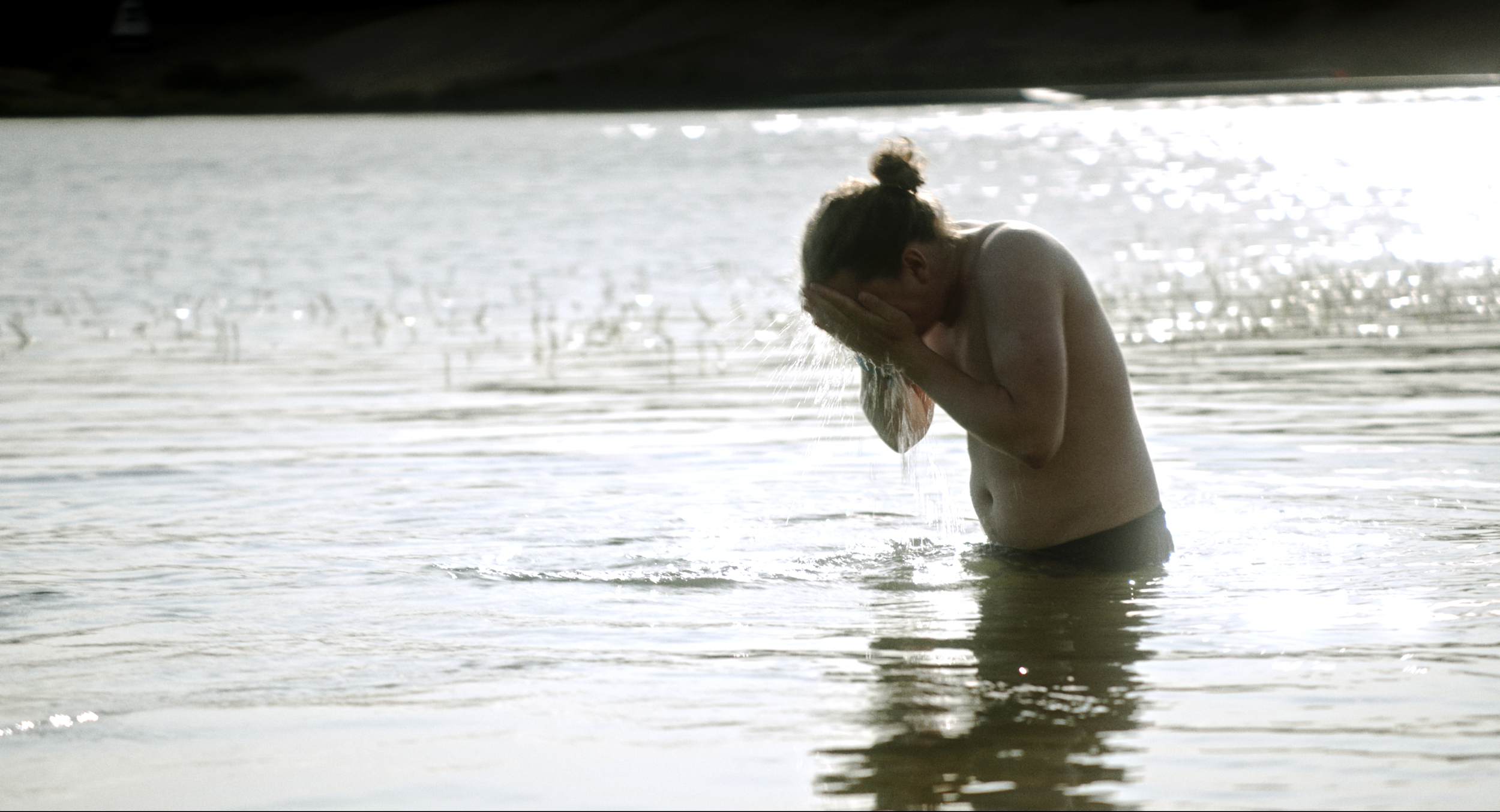 A person is standing in shallow water under a dock or bridge, washing their face with water.