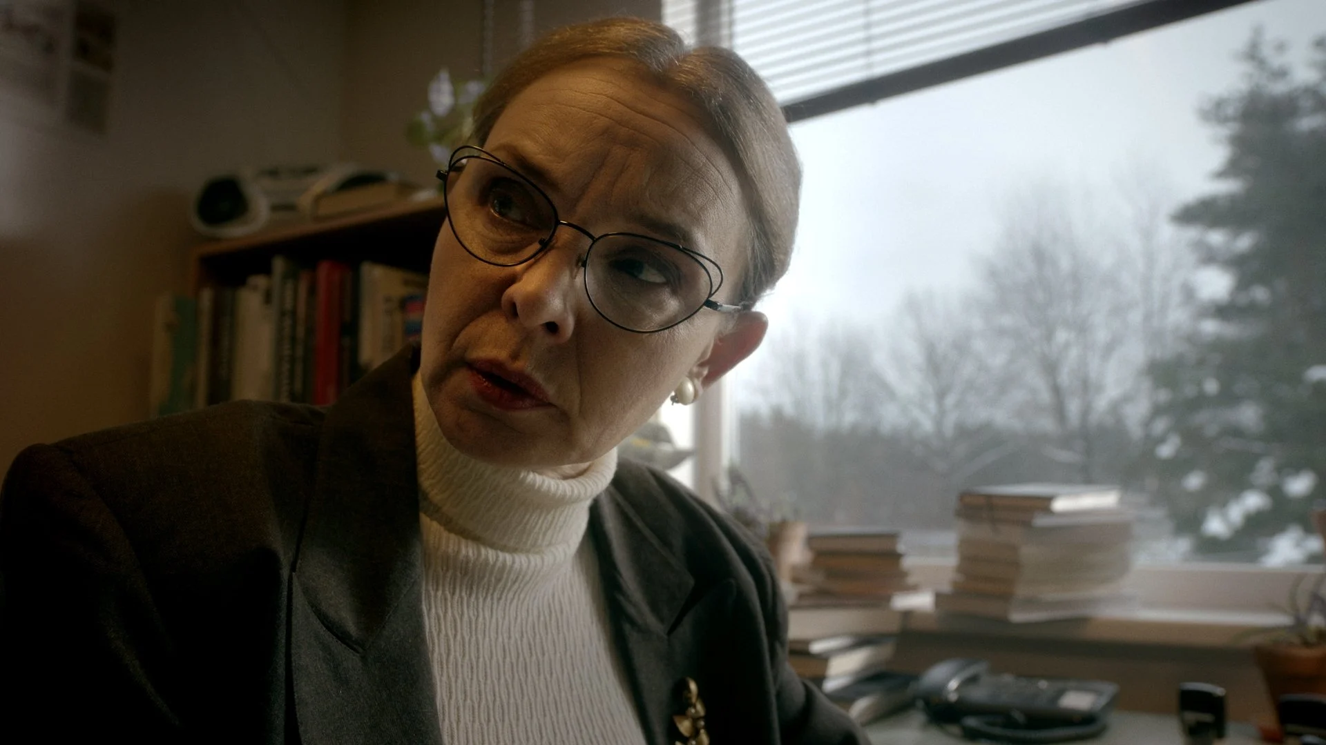 A woman with short gray hair, glasses, and pearl earrings looking to her left in an office. She is wearing a white turtleneck sweater and a brown blazer, with a bookshelf and window with blinds in the background.