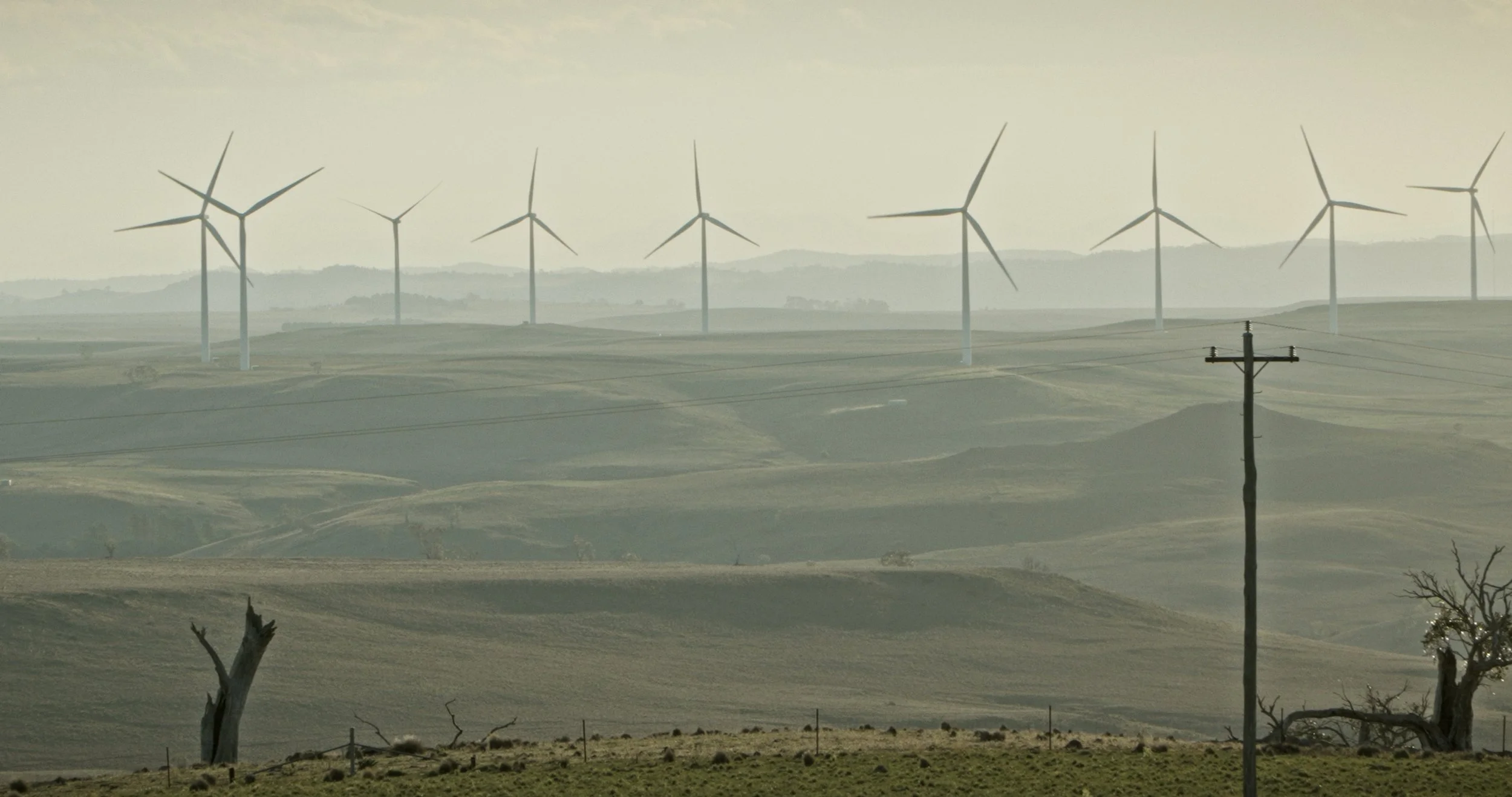 A landscape with rolling hills and wind turbines in the distance, overcast sky, power pole in the foreground.