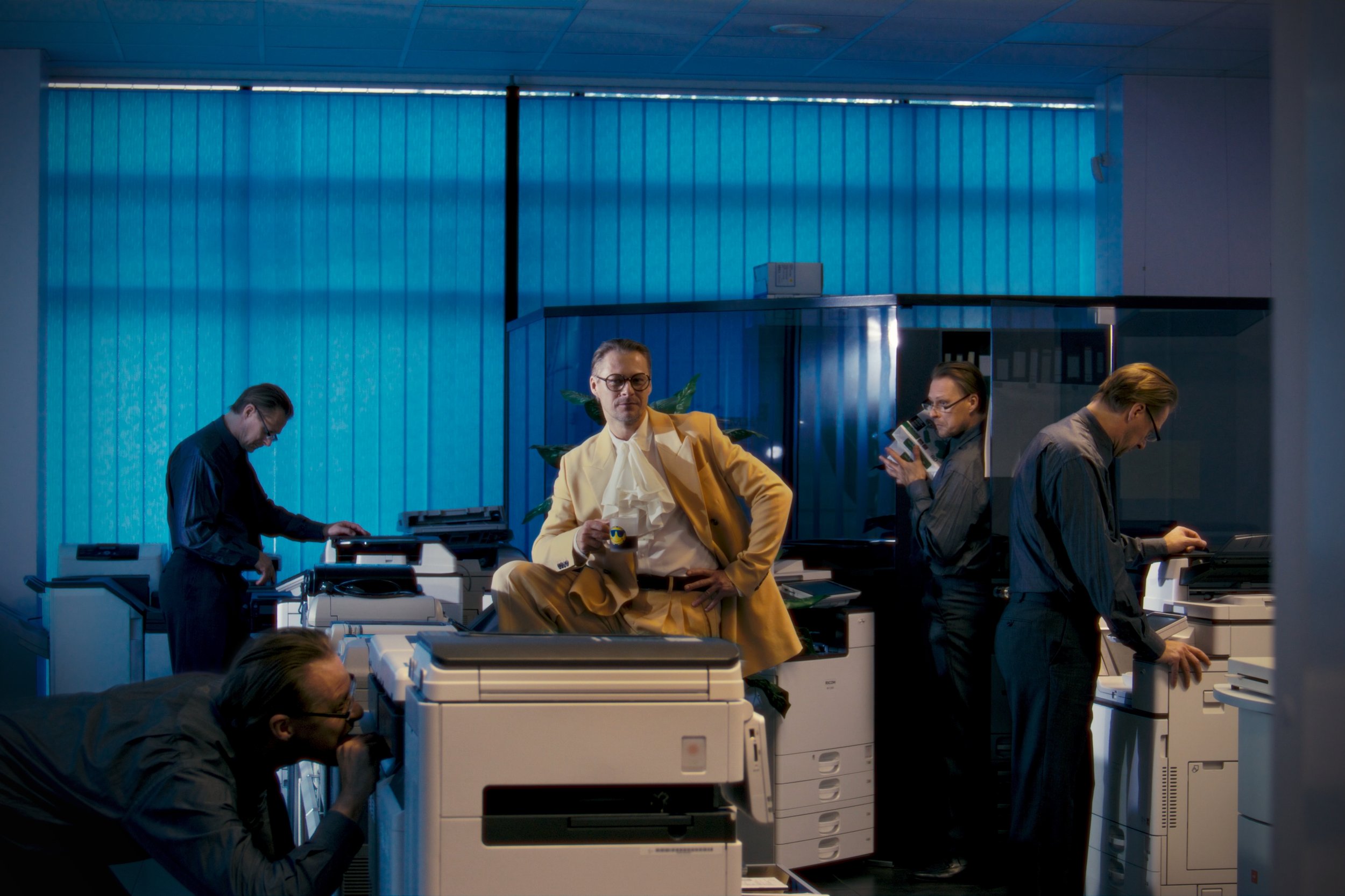A man wearing a yellow suit with glasses holding a mug, sitting at a desk in a modern office with blue wall paneling. Several other people are working with printers and documents around him.