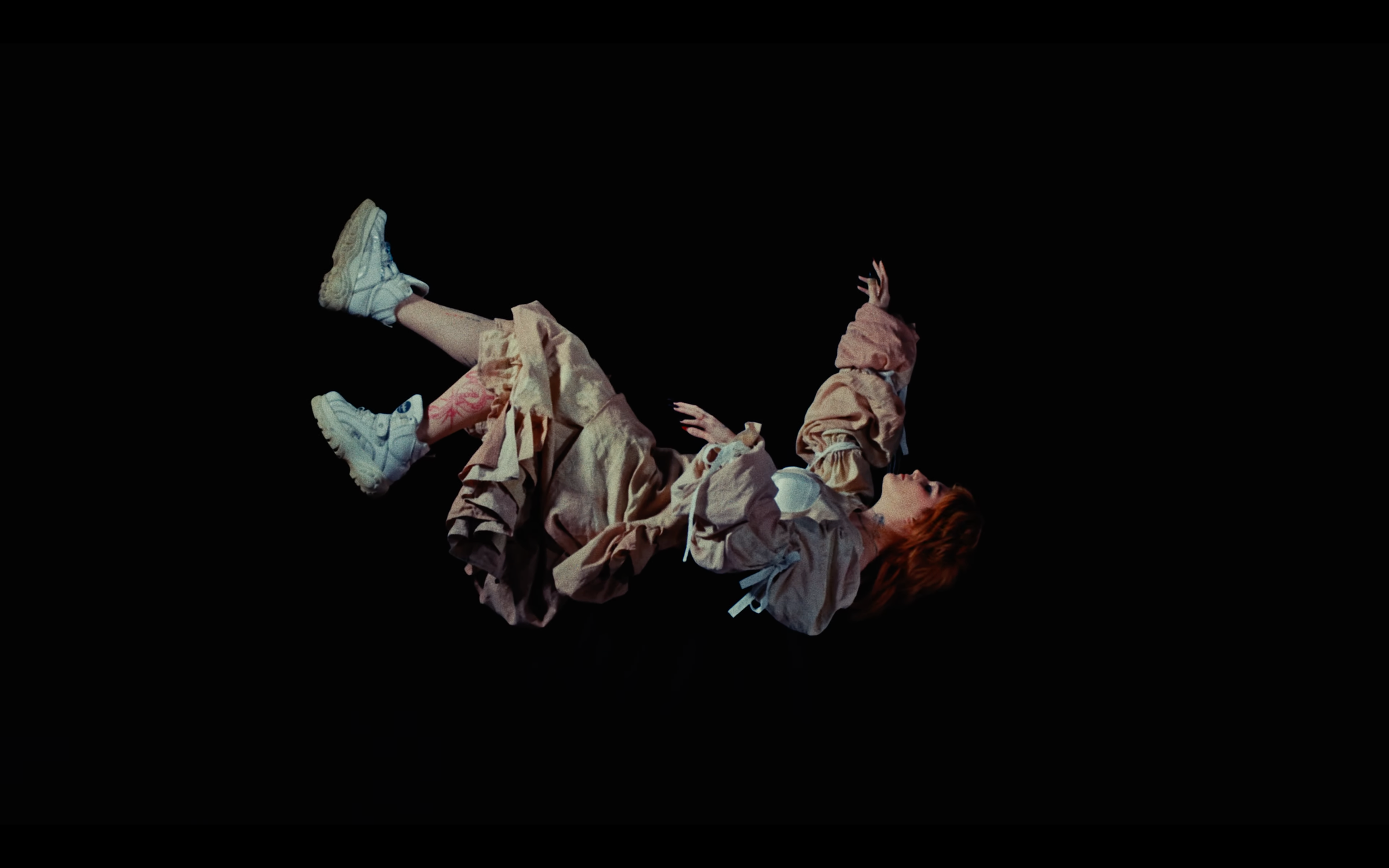 A person with red hair lying on their back against a black background, wearing beige clothes and white sneakers, with arms and legs extended.