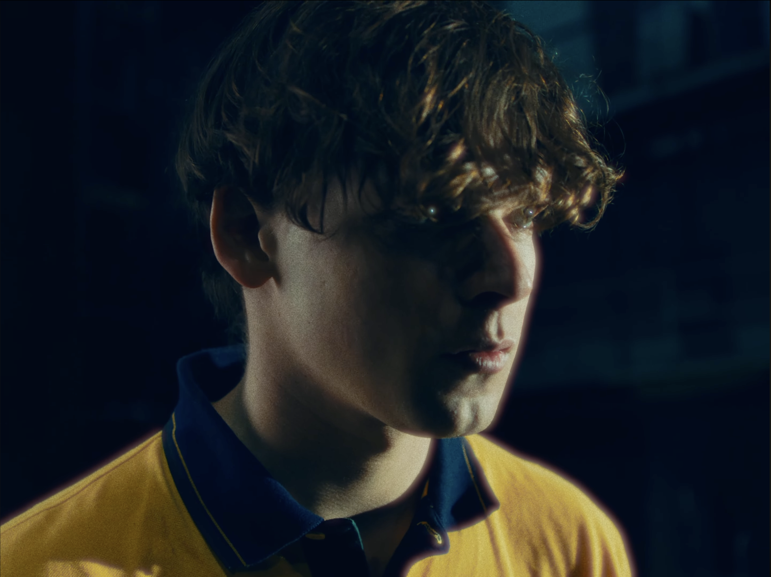 Close-up of a young man with curly brown hair wearing a yellow sports jersey with a blue collar, looking downward in a dark setting.