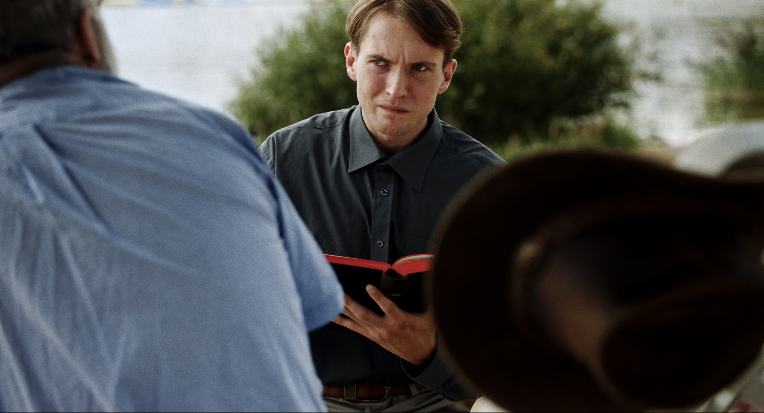 A young man with brown hair and a serious expression is holding a book while talking to two older men, one in a blue shirt and the other with a hat, outdoors near water and greenery.