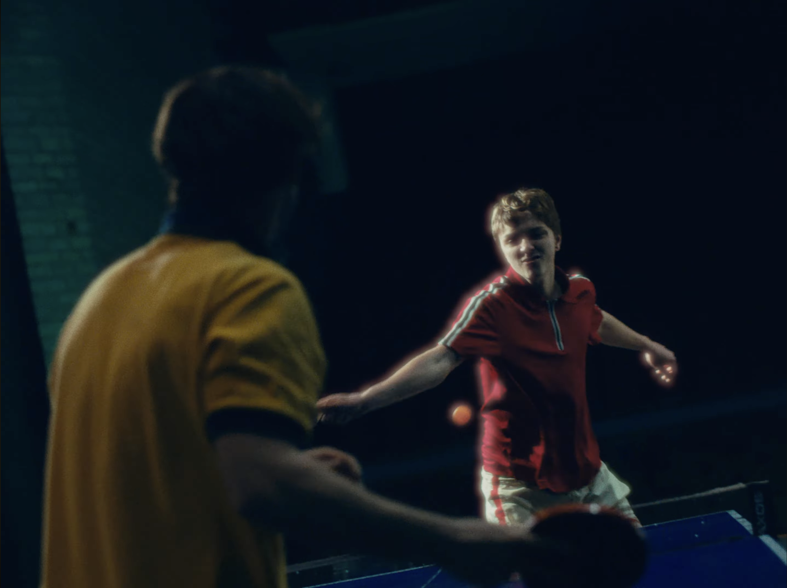 Two young men playing table tennis in a dimly lit indoor setting. The young man on the right is wearing a red and black jacket and appears to be preparing to hit the ball, while the other man is wearing a yellow shirt.
