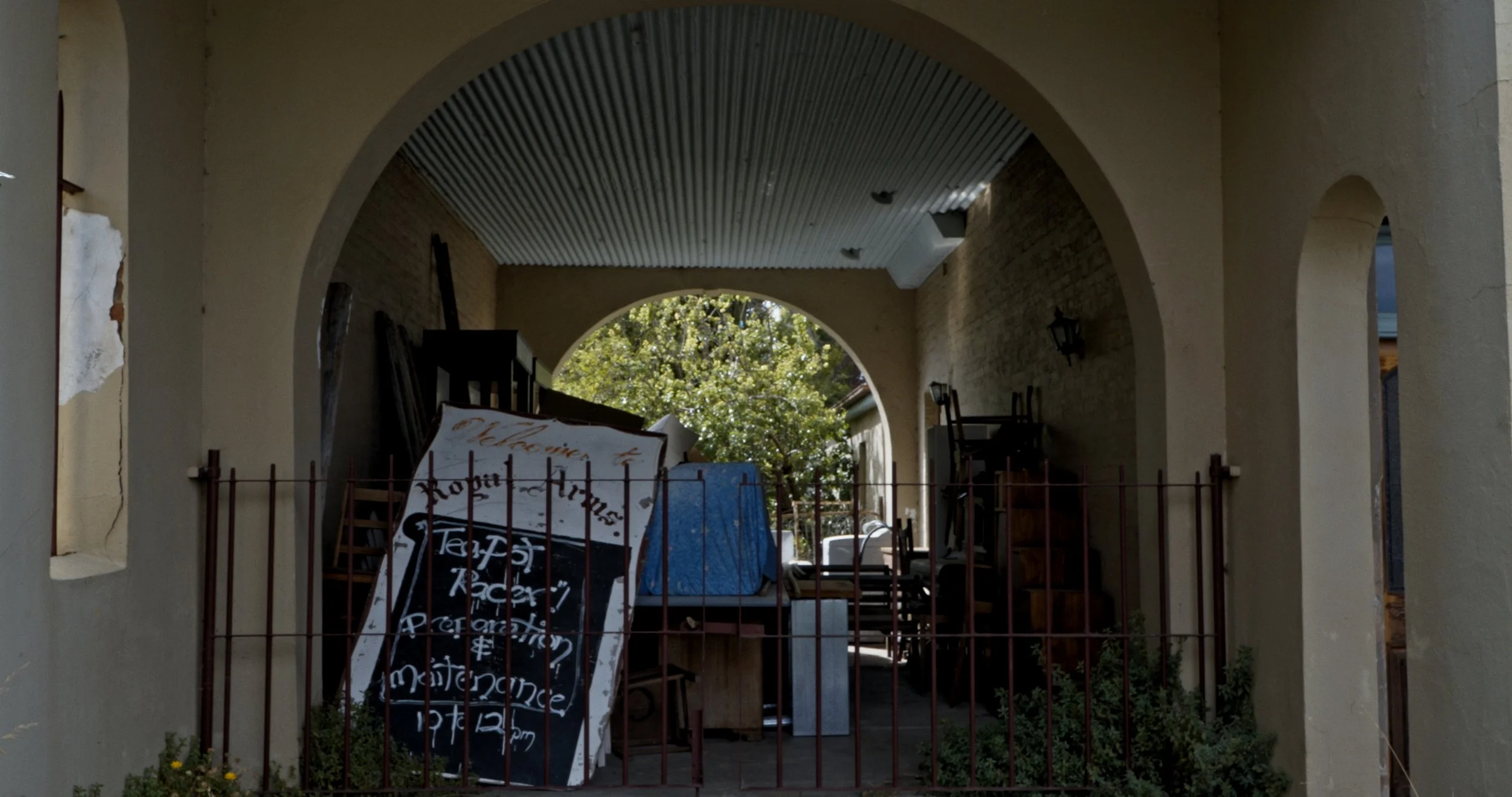 View of a covered porch area with chairs and a small table, seen through an arched opening. There are trees visible in the background, and a signboard with handwritten text leaning against a fence in the front.