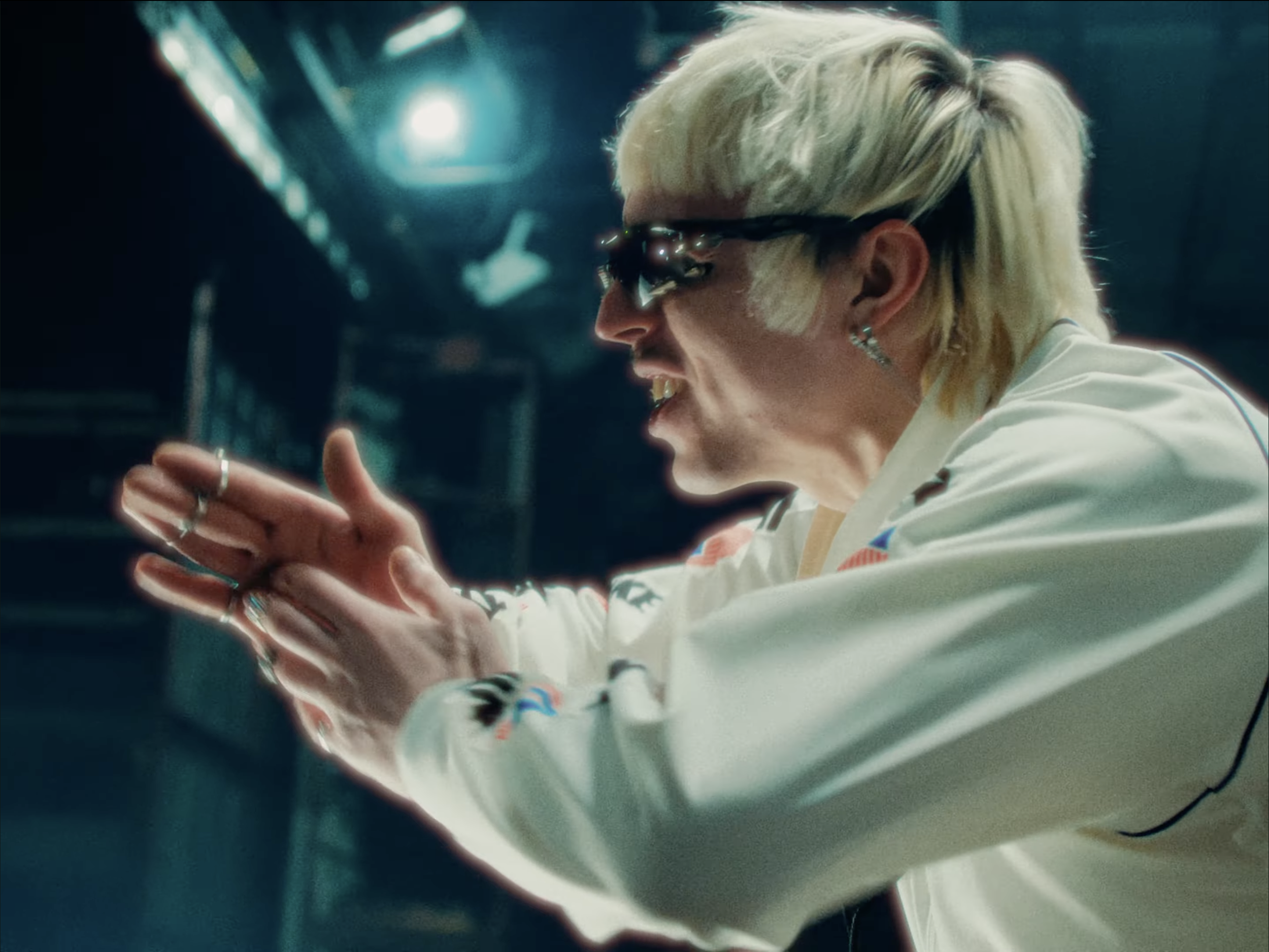 A person with platinum blonde hair wearing sunglasses and a white shirt, engaged in a task or conversation in a high-tech environment with blue lighting.