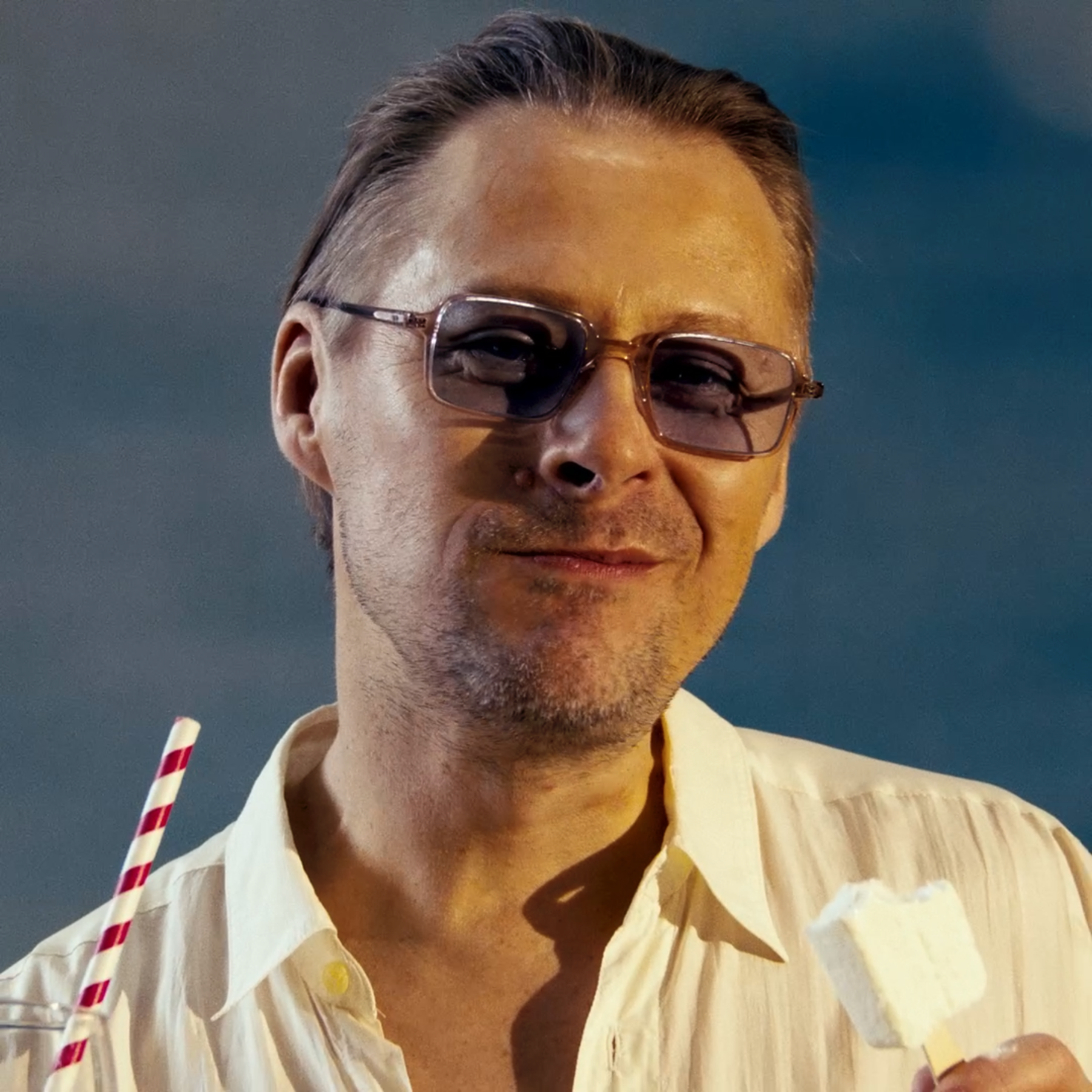 A man wearing sunglasses and a yellow collared shirt, holding a red and white striped straw, against a dark background.