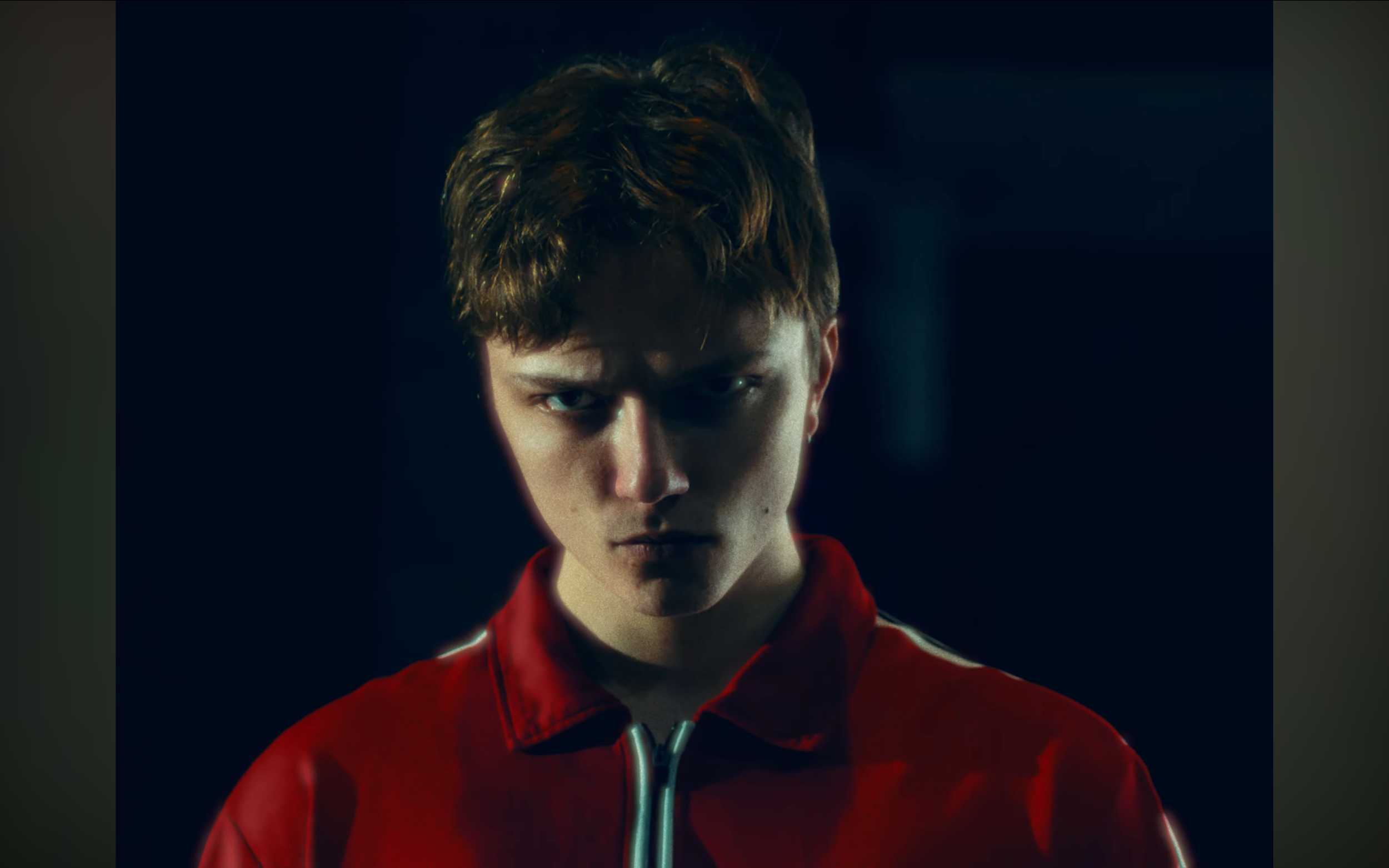 A young man with short brown hair and light skin, wearing a red jacket with a zipper, looking intensely at the camera in a dark setting.