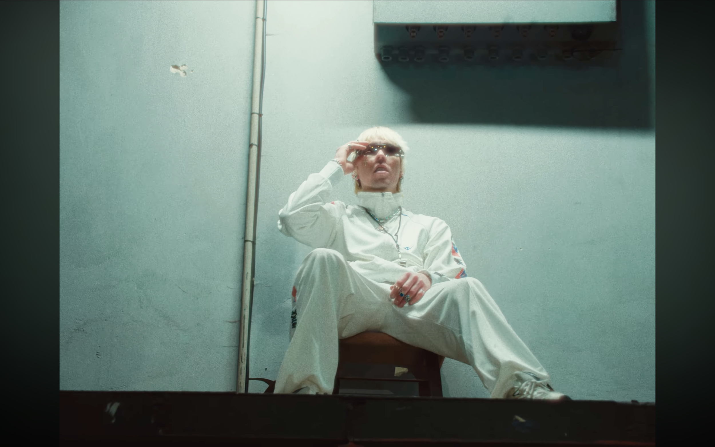 A person with blonde hair and sunglasses sitting on the floor against a wall, wearing a white jacket and pants with colorful accents, looking up and adjusting their sunglasses.
