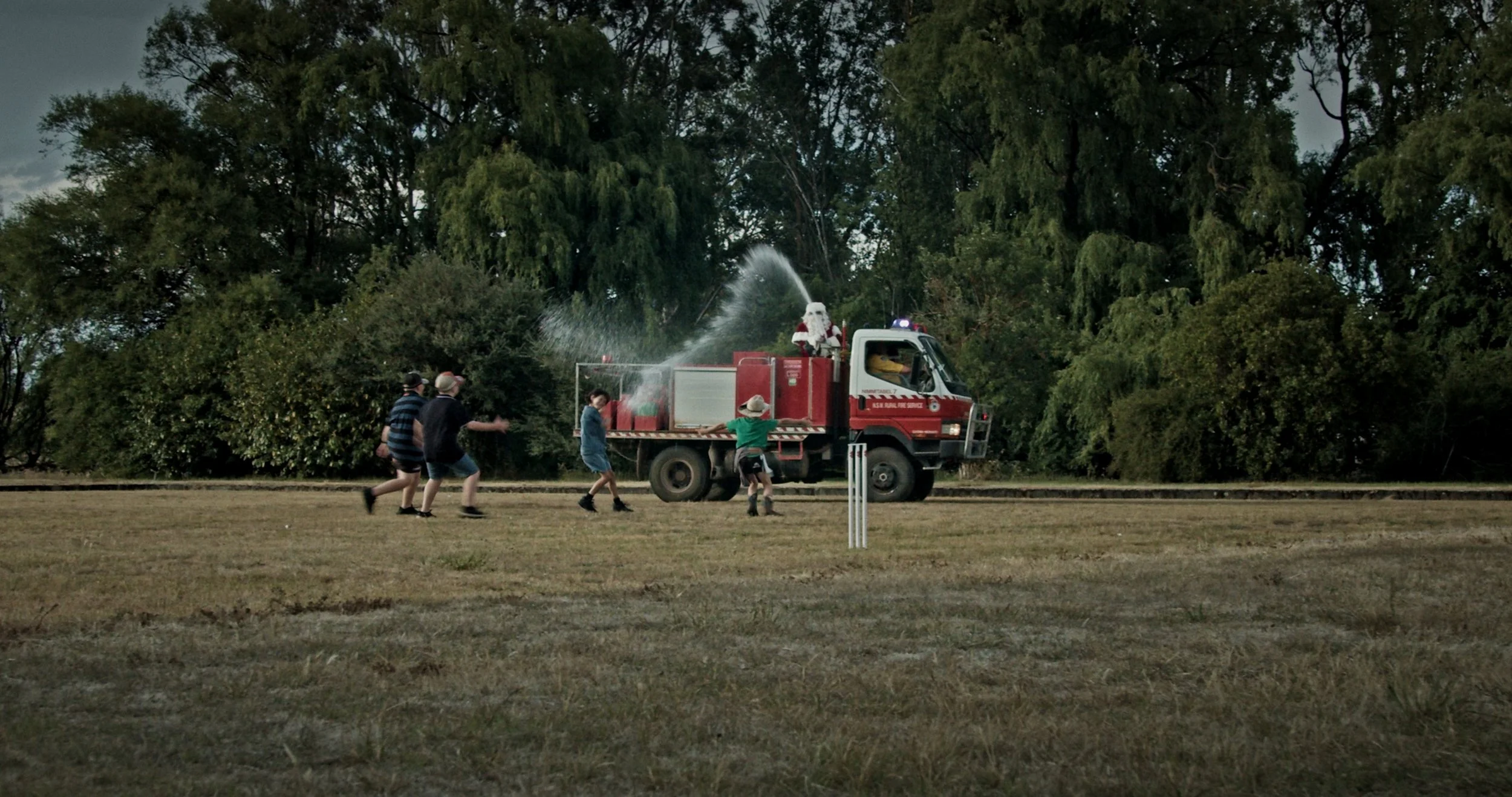 Kids playing around a fire truck with a firefighter on top spraying water, in a park with large trees in the background.