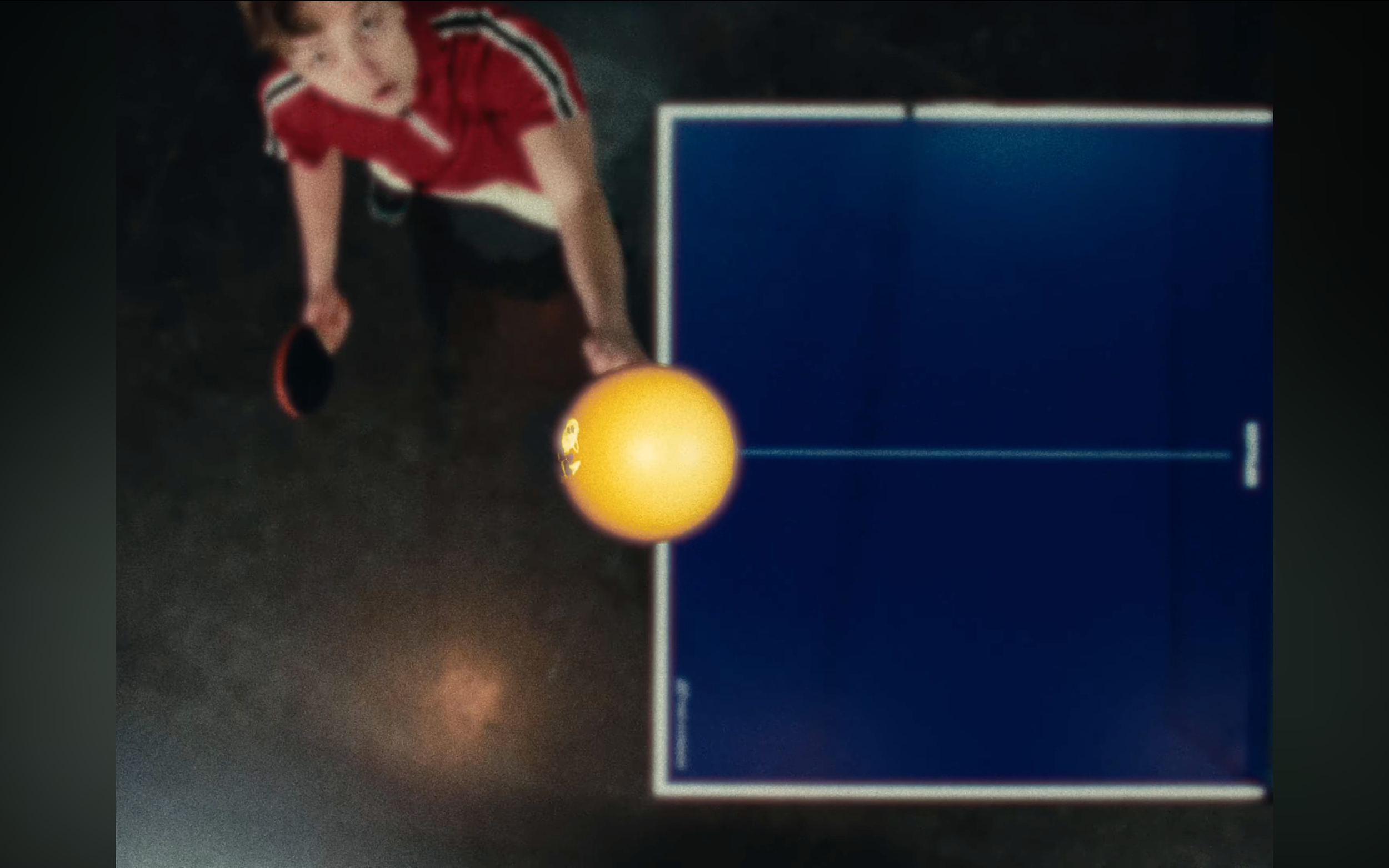 A person playing ping pong with a yellow ball and a red paddle, wearing a red shirt with white and black stripes, next to a blue ping pong table.